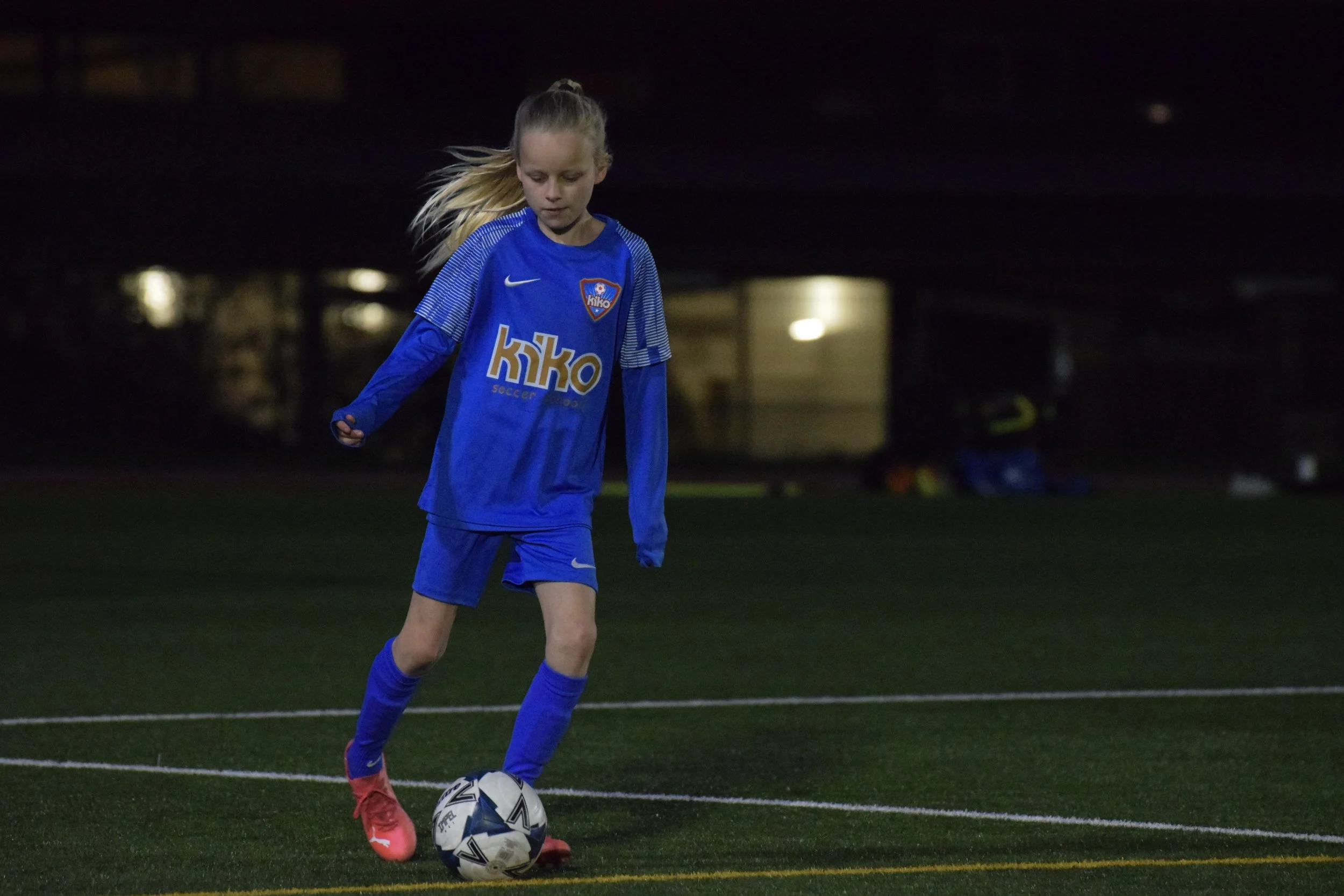 A young girl in a blue soccer uniform practicing on a soccer field at night, preparing to kick a soccer ball.