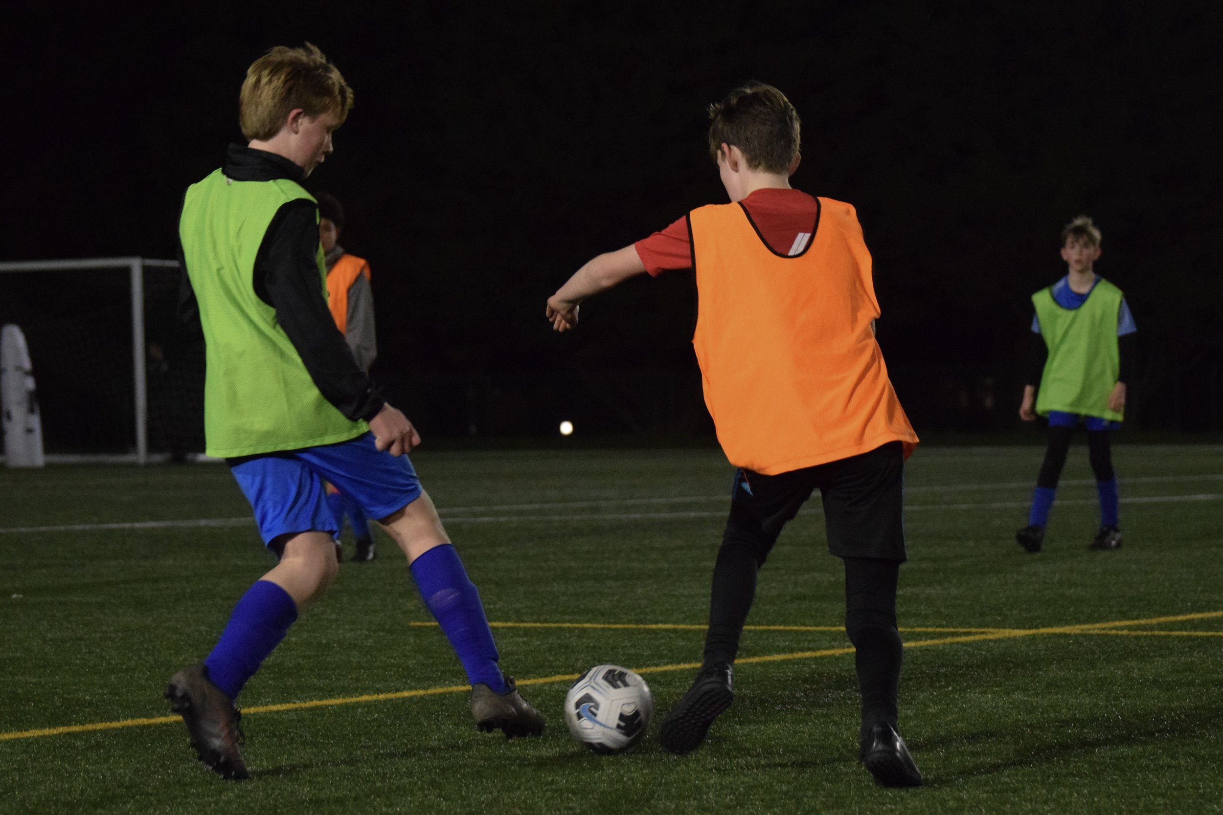Two boys playing soccer at night on a field, wearing orange and green vests, with other players in the background.