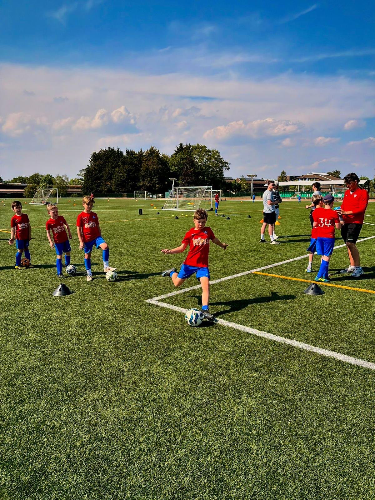 Children in red soccer jerseys practicing dribbling on a green field with cones, with coaches overseeing, under a partly cloudy sky.