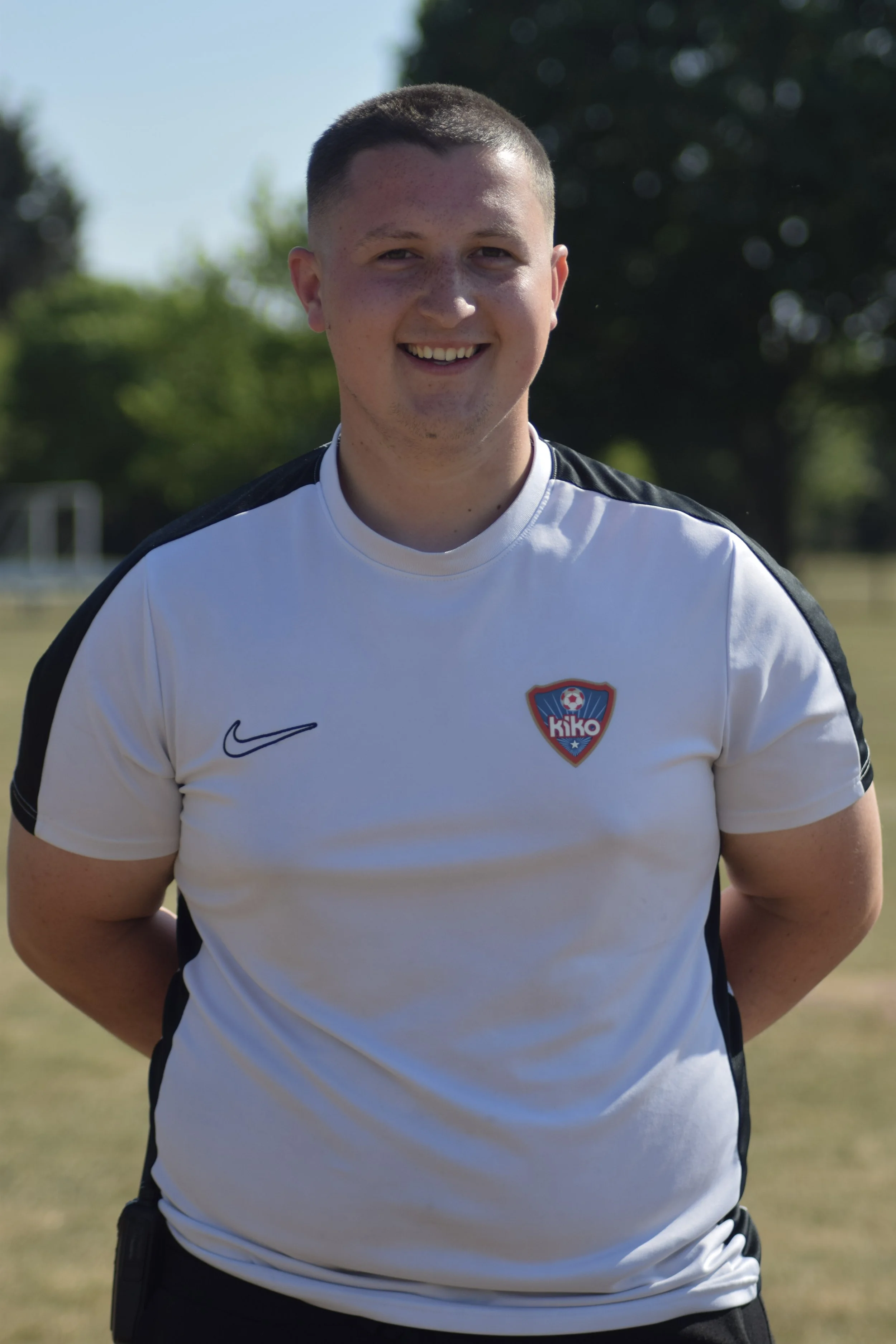 A young man smiling outdoors, wearing a white sports jersey with black accents and a logo on the chest.