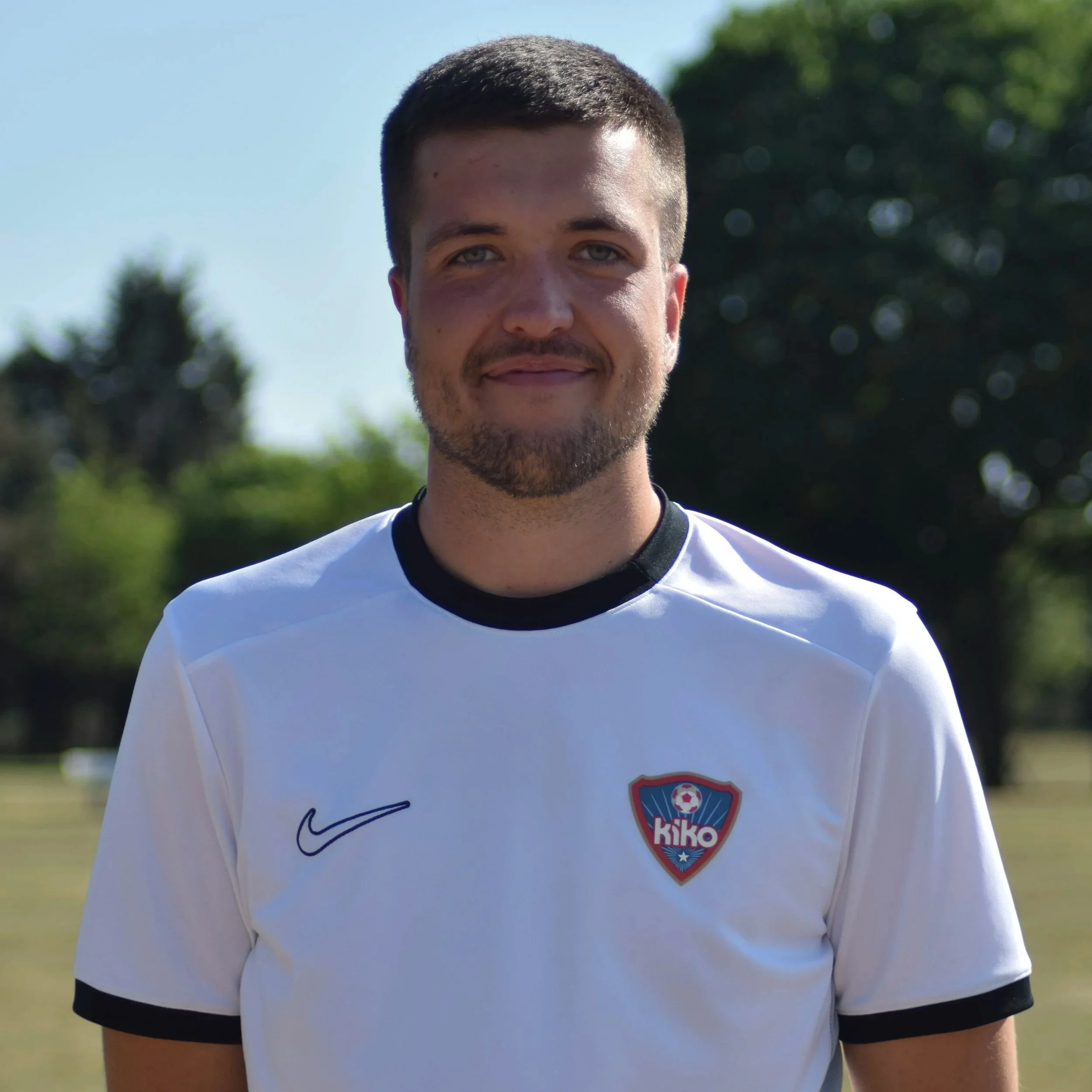 Close-up of a young man with short dark hair and light skin, smiling outdoors in sunlight, wearing a white sports jersey with black trim, and an emblem on the chest featuring a soccer ball and the word "Kiko."