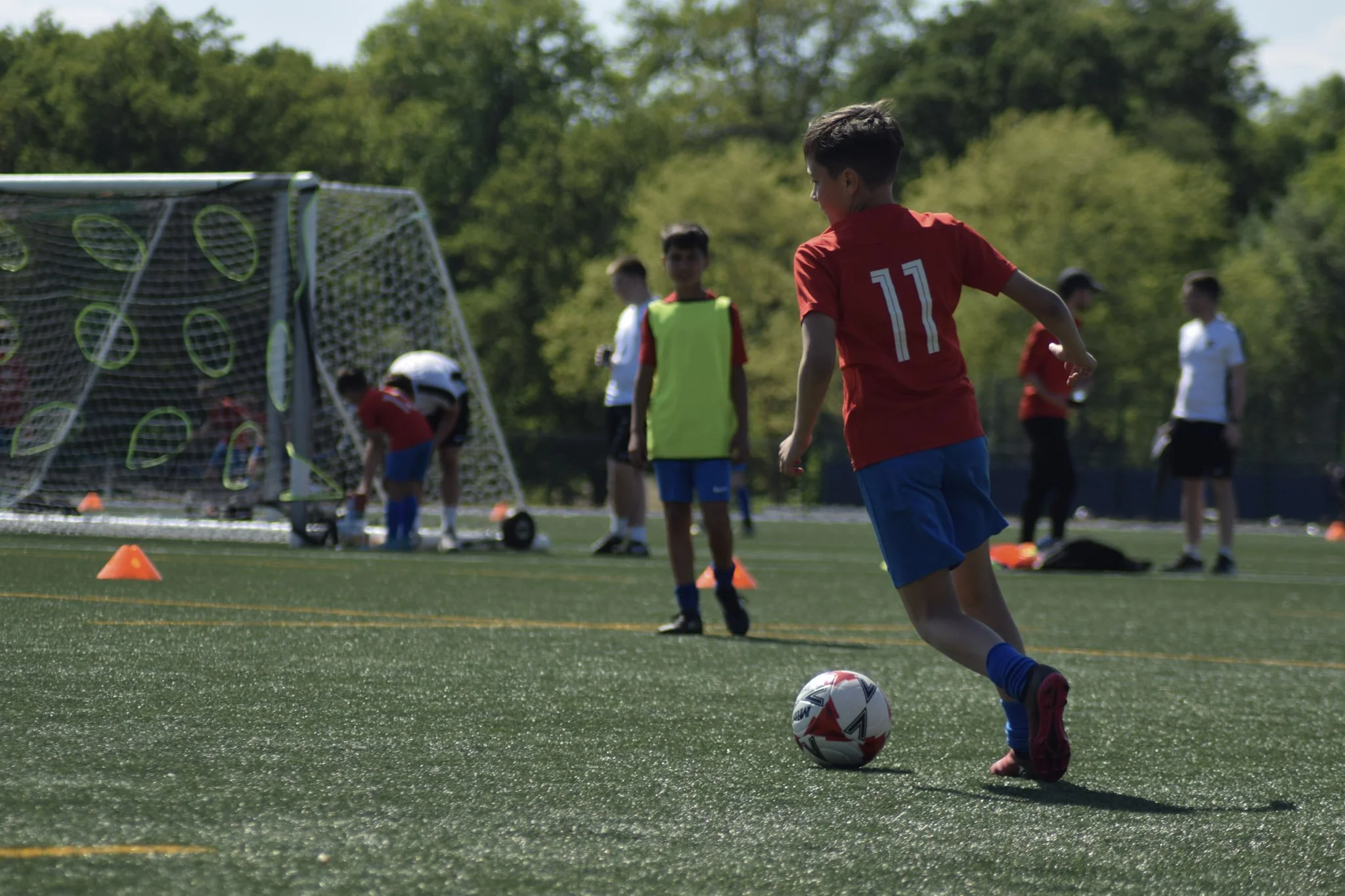 A group of young boys playing soccer on a green field during daytime, with one boy in red kicking a soccer ball near a goalpost, surrounded by trees and orange cones.