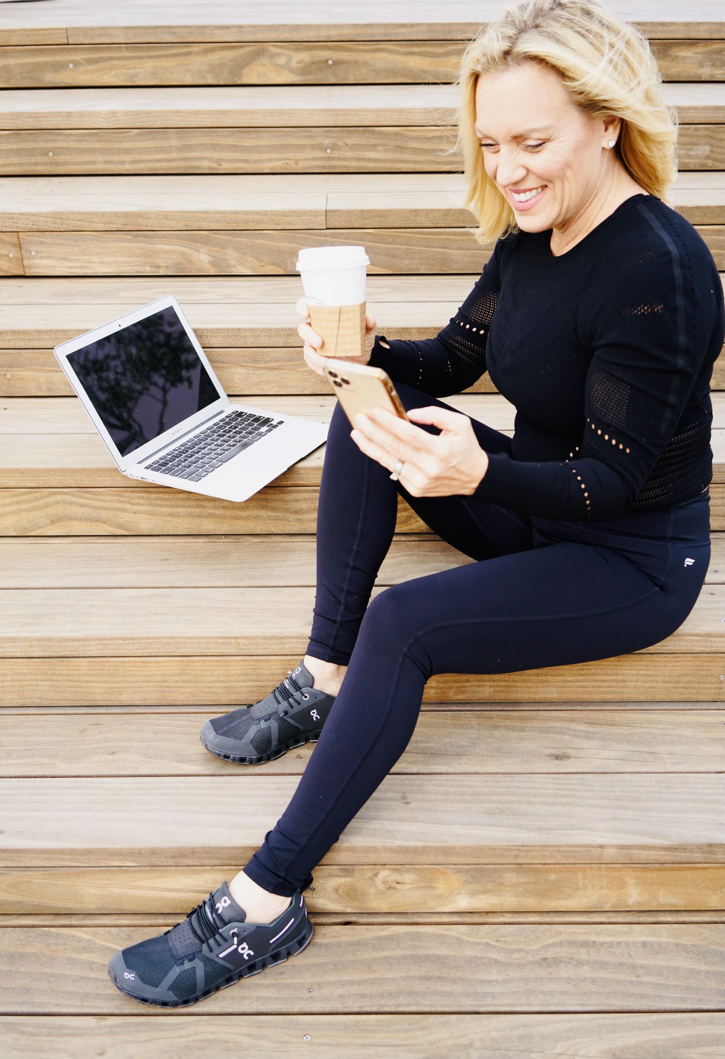 A woman sitting on wooden steps, smiling and looking at her phone, with a laptop and a coffee cup nearby.