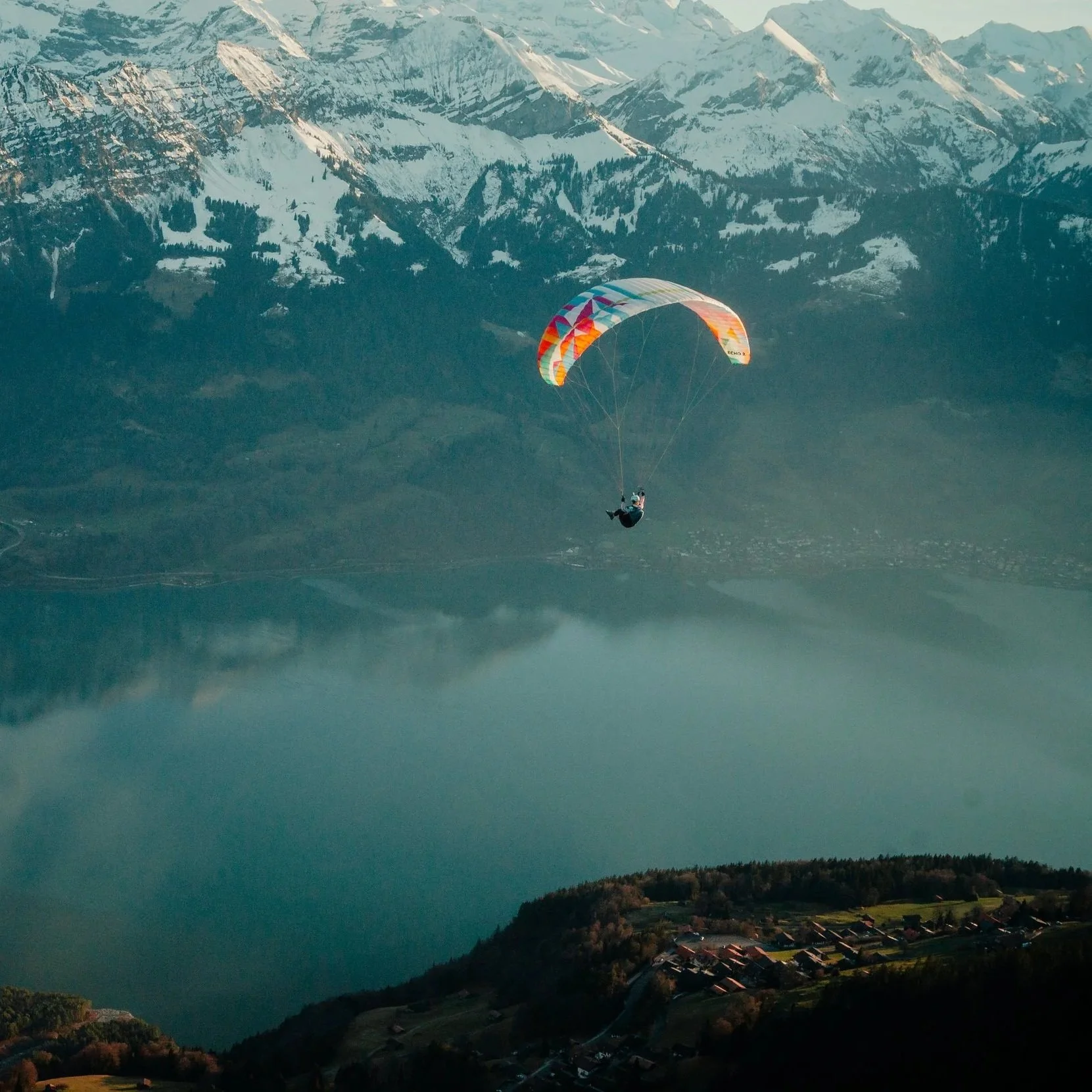 Ein Paragleiter fliegt über einen See vor einer Bergkulisse mit schneebedeckten Gipfeln in den Bergen.