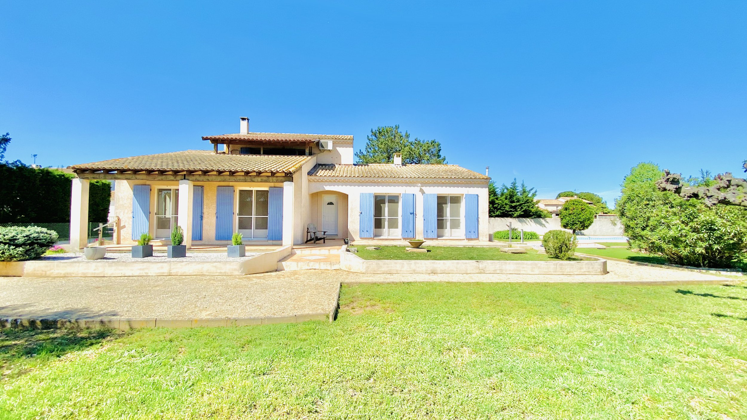 Front view of a house with blue shutters, a patio with a small bench, and a well-maintained lawn with some plants and trees under a bright blue sky.