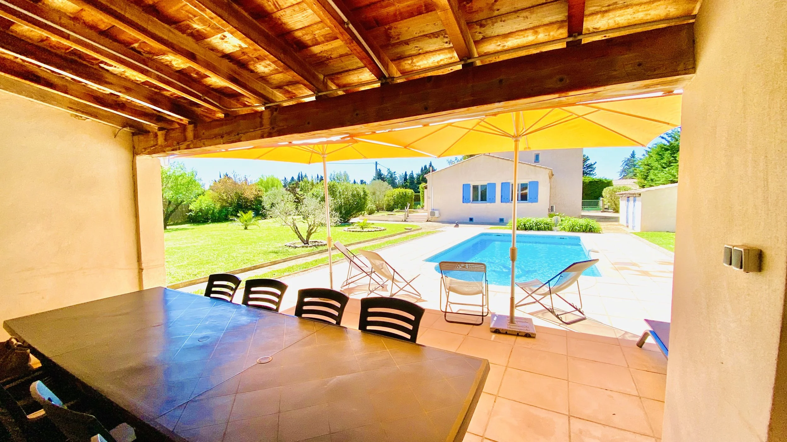 Covered patio with a large table and chairs, overlooking a backyard with a swimming pool, lounge chairs, yellow umbrellas, and green yard with trees and a white house with blue shutters.