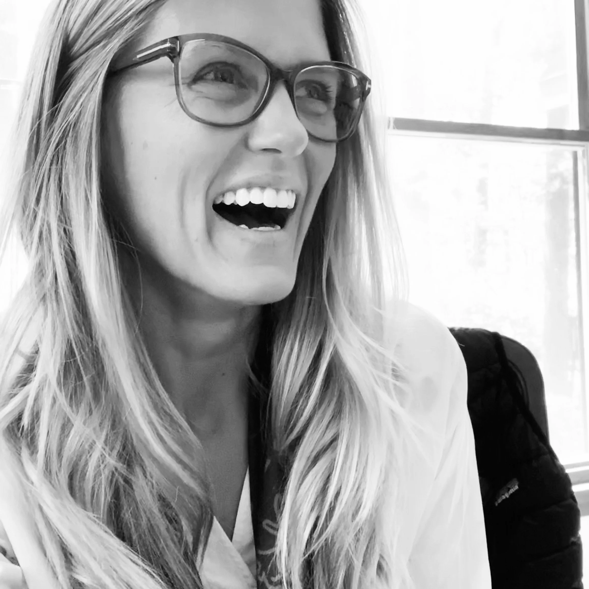 A woman with long hair wearing glasses, smiling with her teeth showing, in a black and white photo.