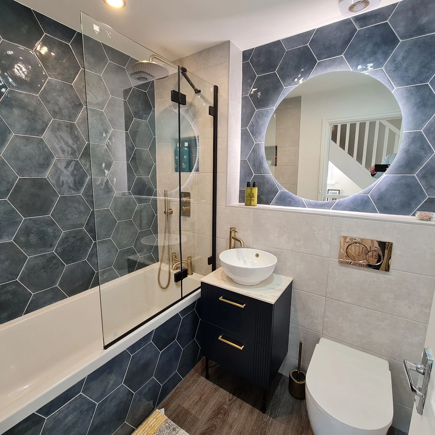 Modern bathroom with hexagon tiles on wall, glass shower enclosure with gold fixtures, round vessel sink on a black vanity, and a large circular mirror with back lighting.