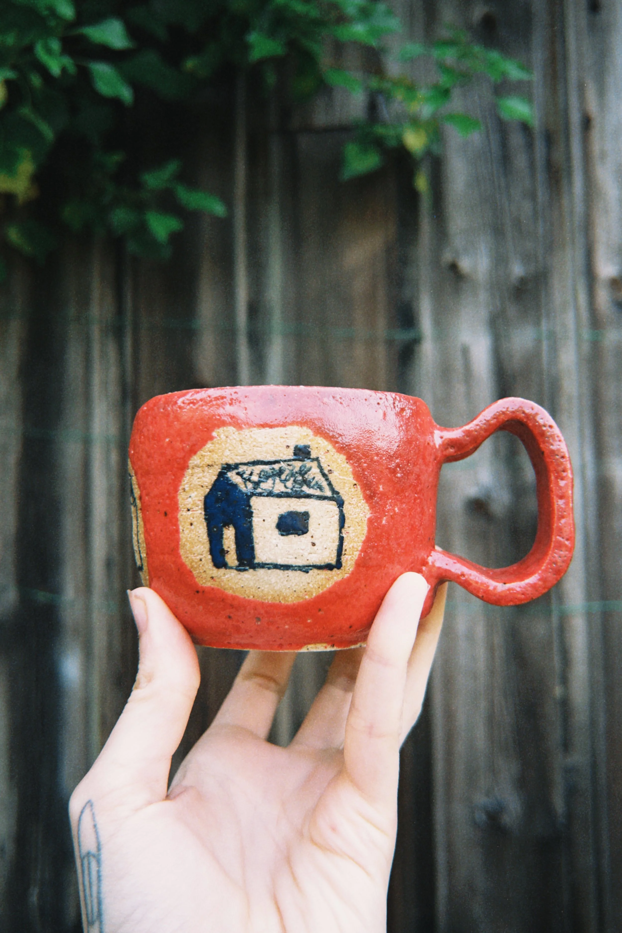 Person holding a red ceramic mug with a black house illustration, standing in front of a wooden fence with green foliage.