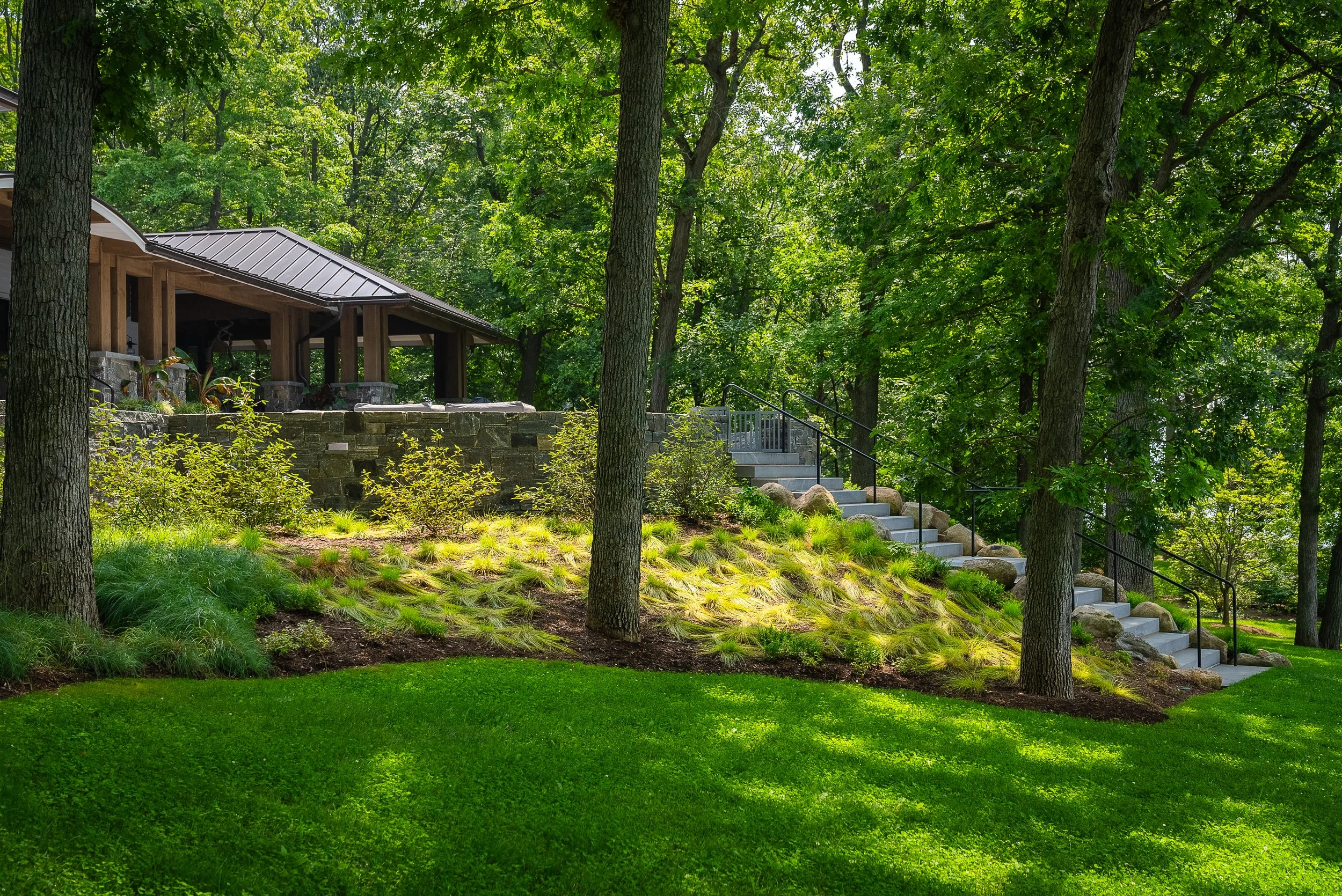 Lush green backyard with a wooden porch, stairs, and landscaped plants surrounded by tall trees.