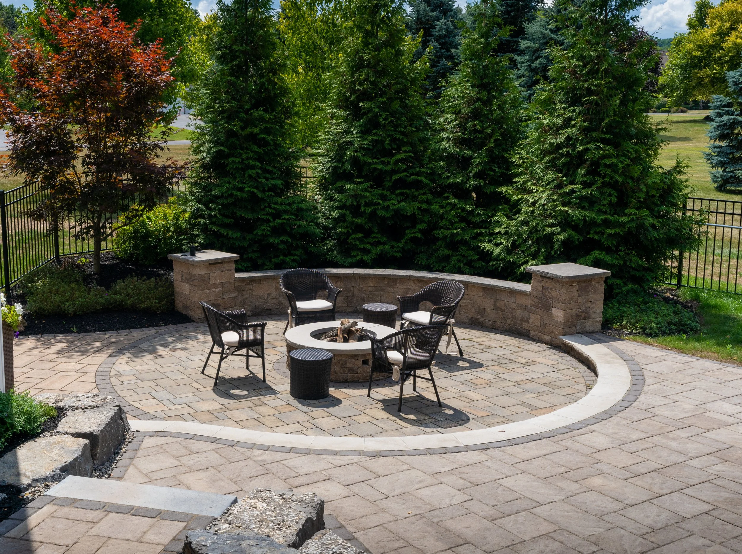 Outdoor patio with stone fire pit, surrounded by wicker chairs and well-manicured greenery.