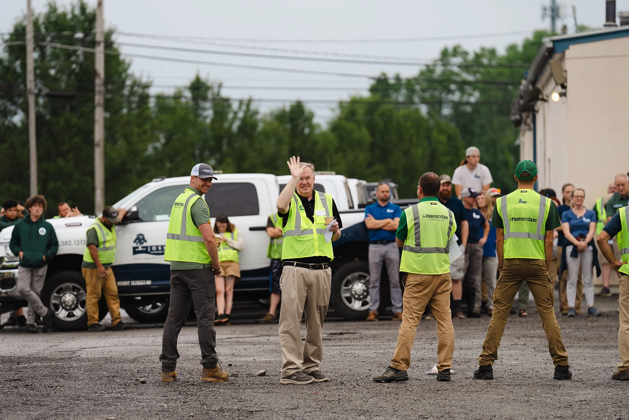 Group of people gathered outdoors, with some wearing yellow safety vests, and one person waving. There are vehicles and trees in the background.