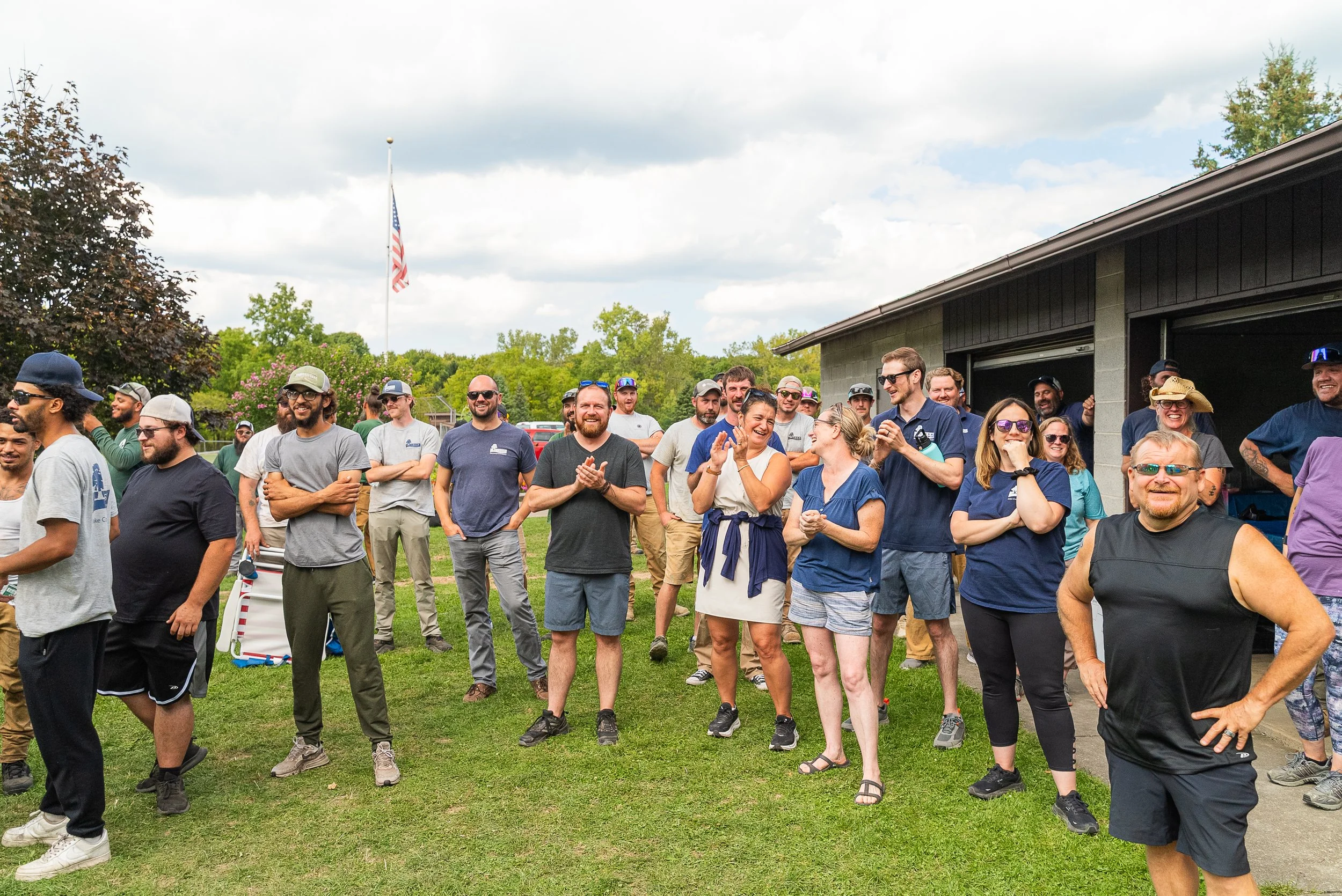 Group of people standing outdoors on a grassy area, smiling and chatting during a gathering or event with overcast skies and a flagpole with an American flag in the background.