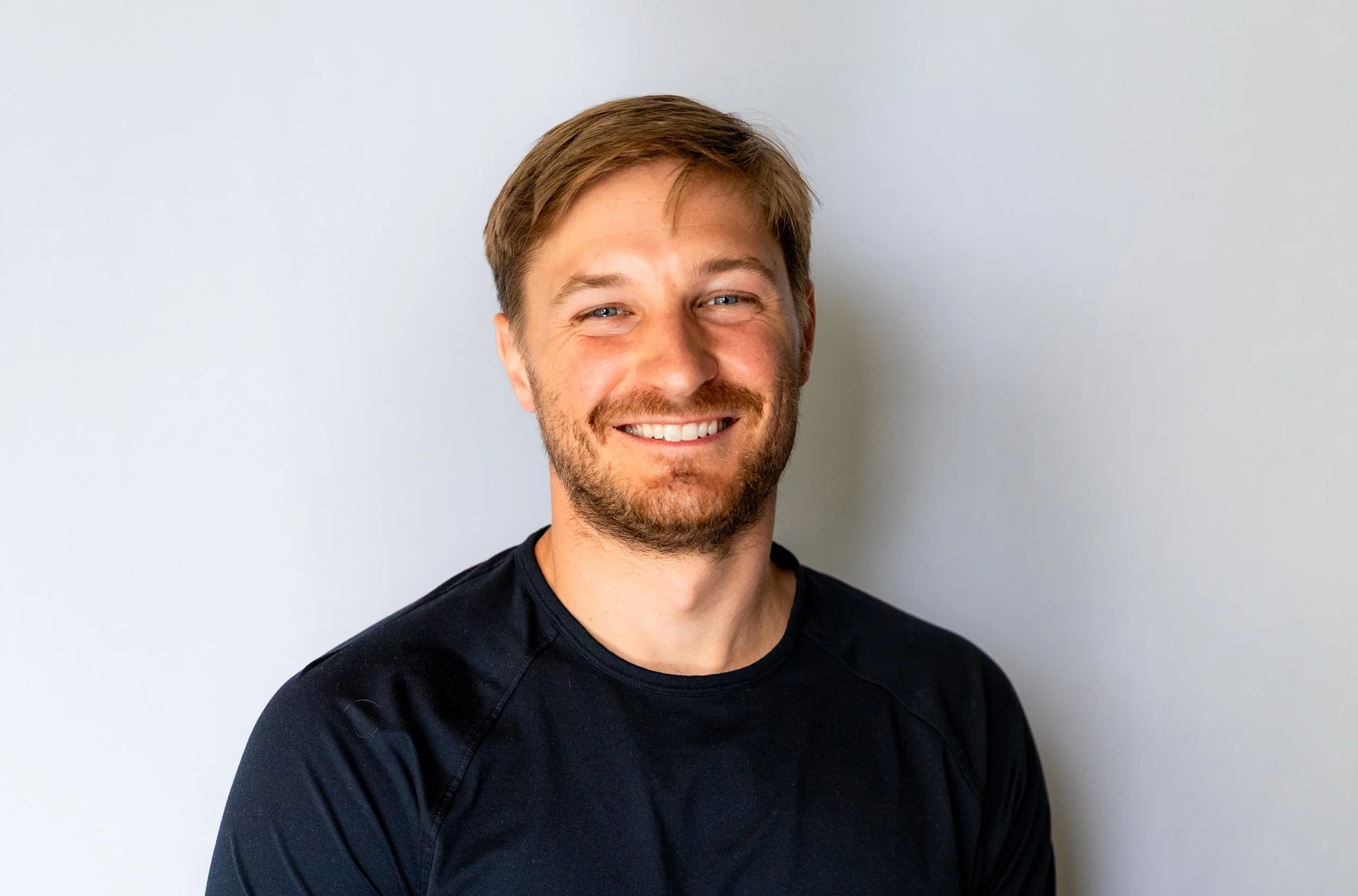 A smiling man with light brown hair and a beard, wearing a black shirt, standing against a plain white wall.