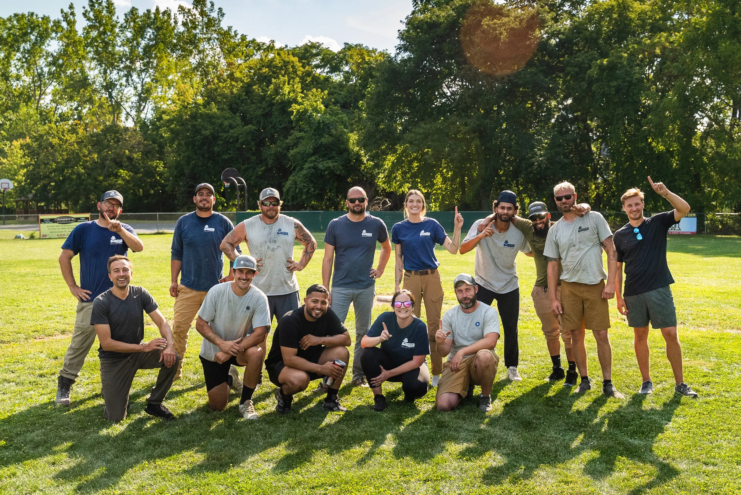 Group of twenty people posing on a grassy field with trees and a basketball court in the background on a sunny day.