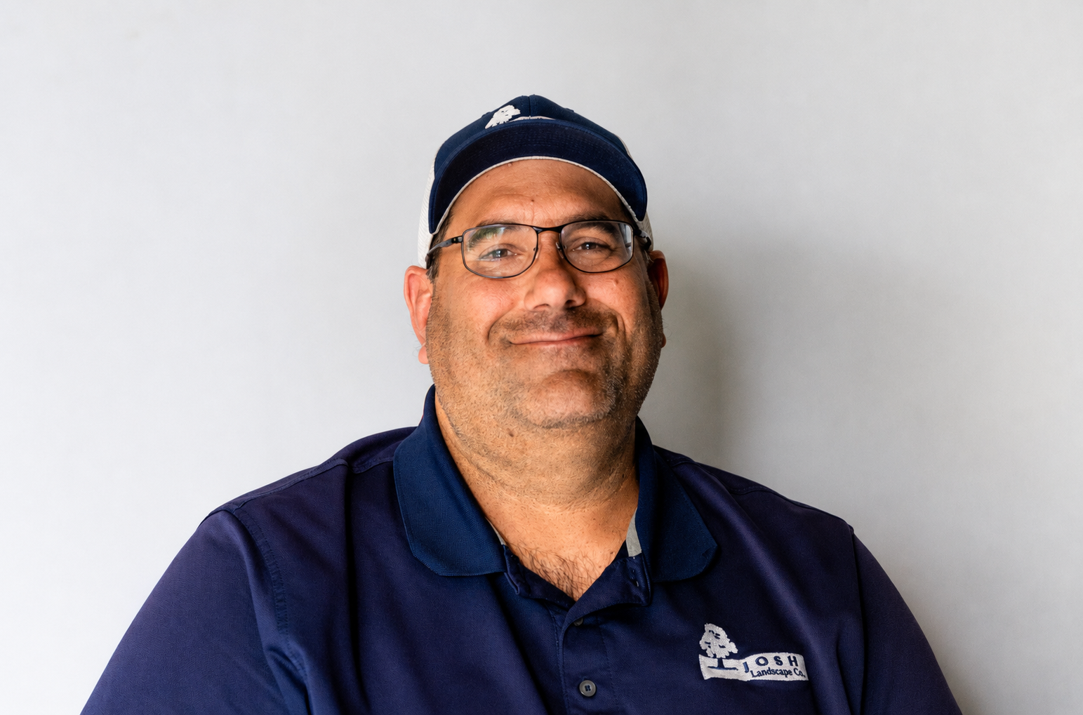 A smiling man wearing glasses, a dark blue cap, and a navy blue polo shirt with a company logo that says 'JOSH Landscaping Co.' against a plain light gray background.