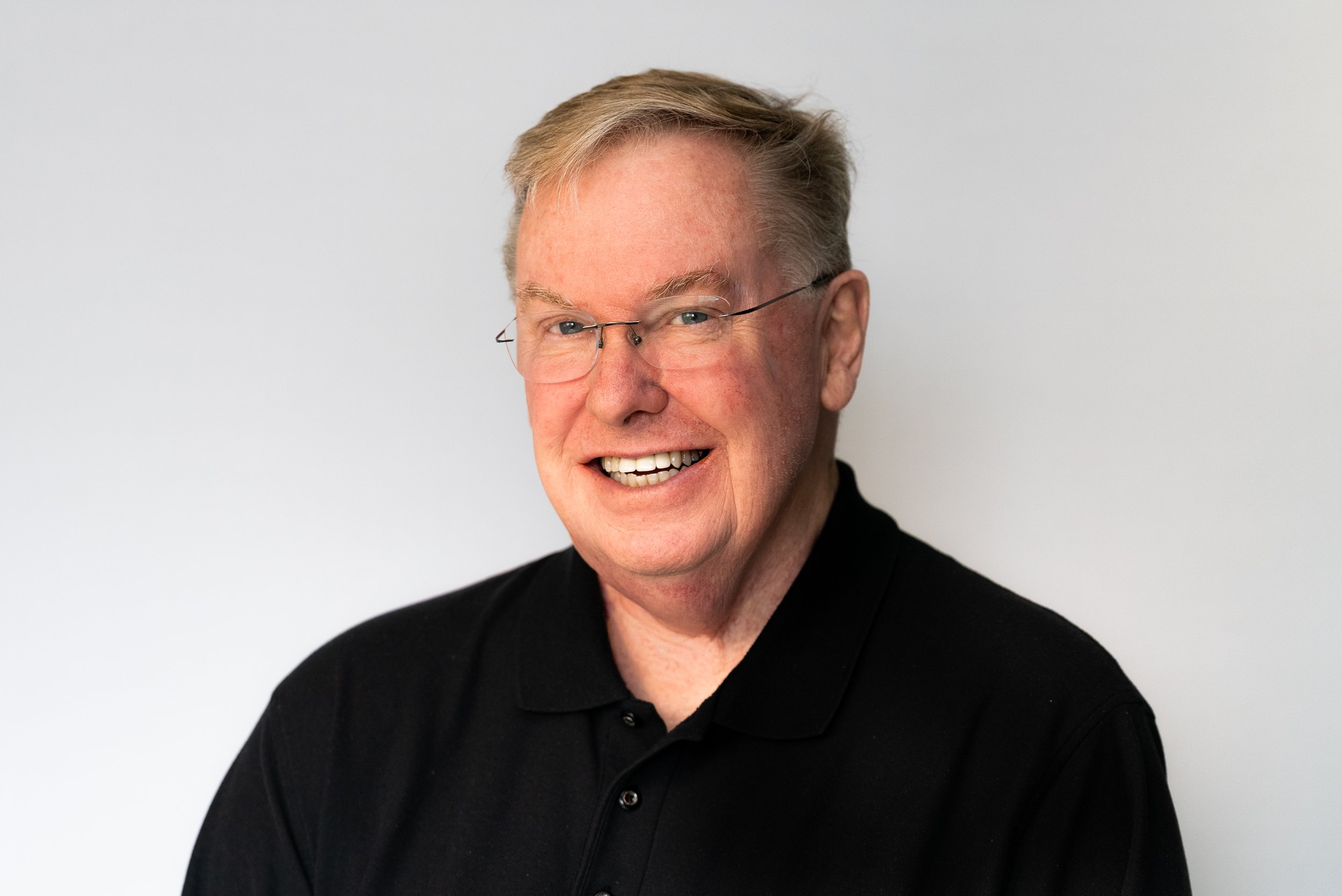 A smiling man with gray hair, glasses, and fair skin wearing a black shirt, posing against a plain light gray background.