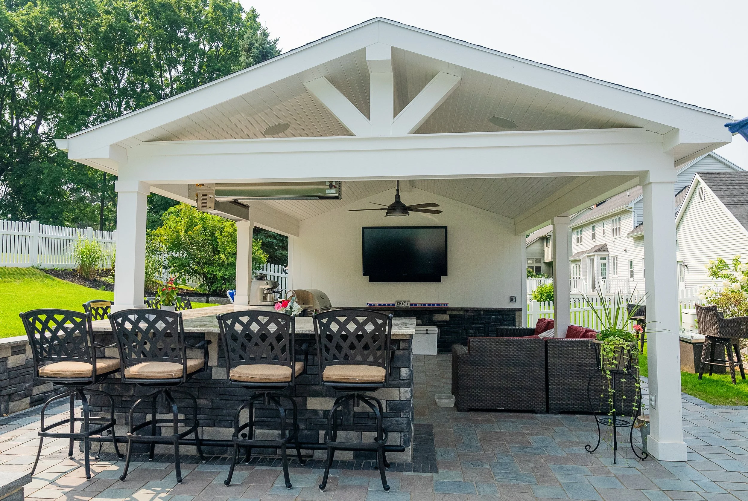 A small beige shed with double glass doors, two windows, and a white overhead garage door. It has a black metal roof and a partially constructed porch with wooden beams. The area in front is paved with stone tiles, and there is a dirt area with some hay bales and a black compost bin nearby. A new wooden fence is being installed on one side, and the background features leafless trees and a partly cloudy sky.