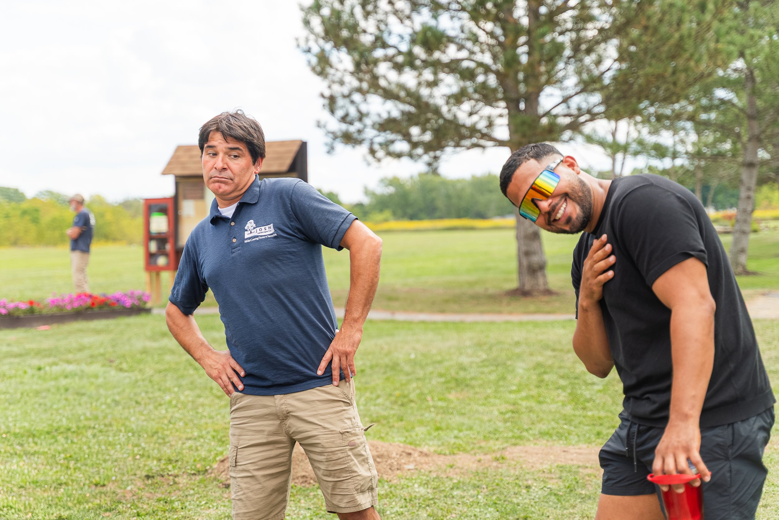 Two men standing outdoors on a grassy area, one with a serious expression and the other smiling while wearing rainbow sunglasses, with trees and a person in the background.