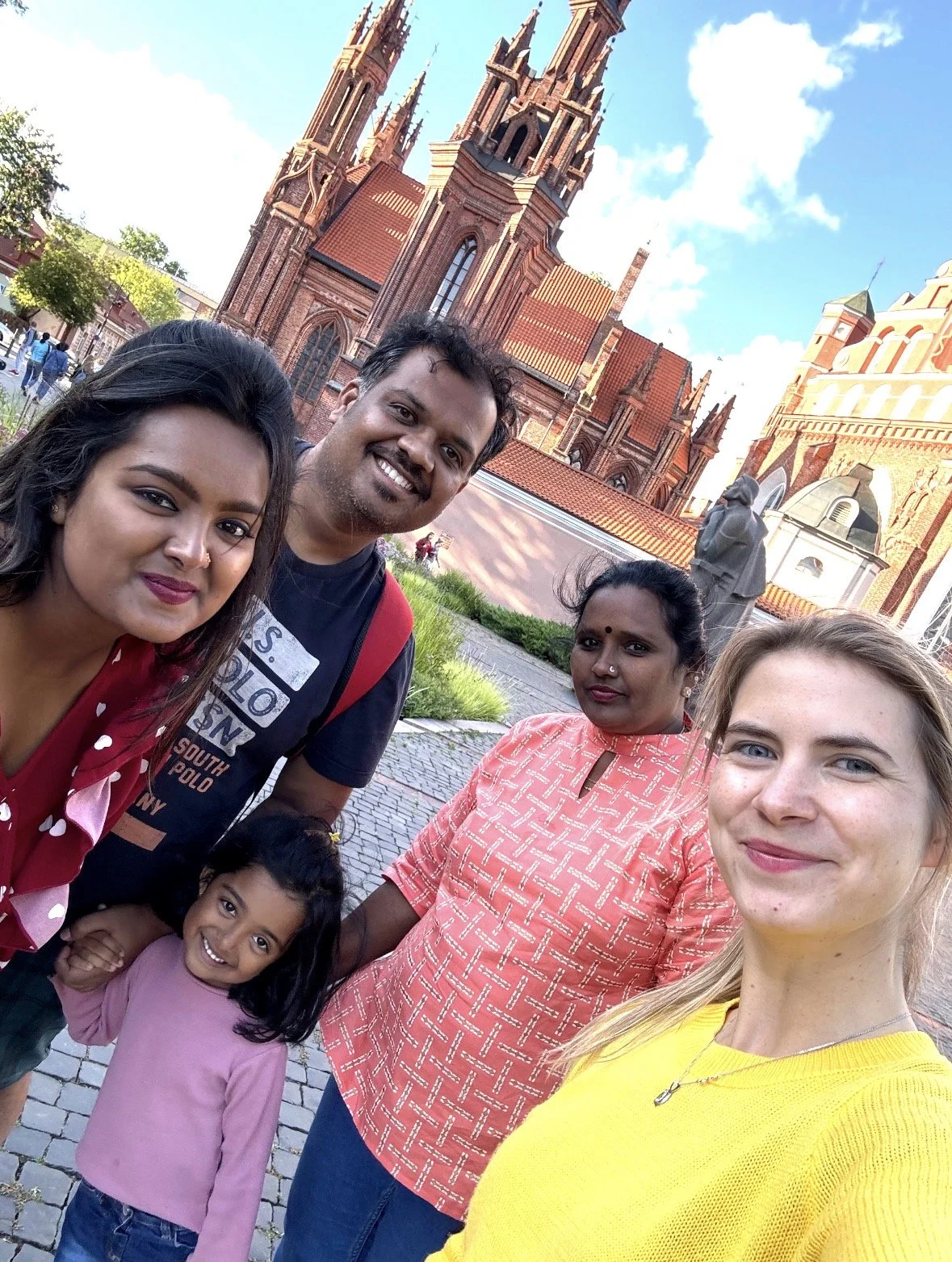 Group of five people, including a child, smiling for a selfie in front of a historic brick church with tall spires and statues, under a blue sky with some clouds.