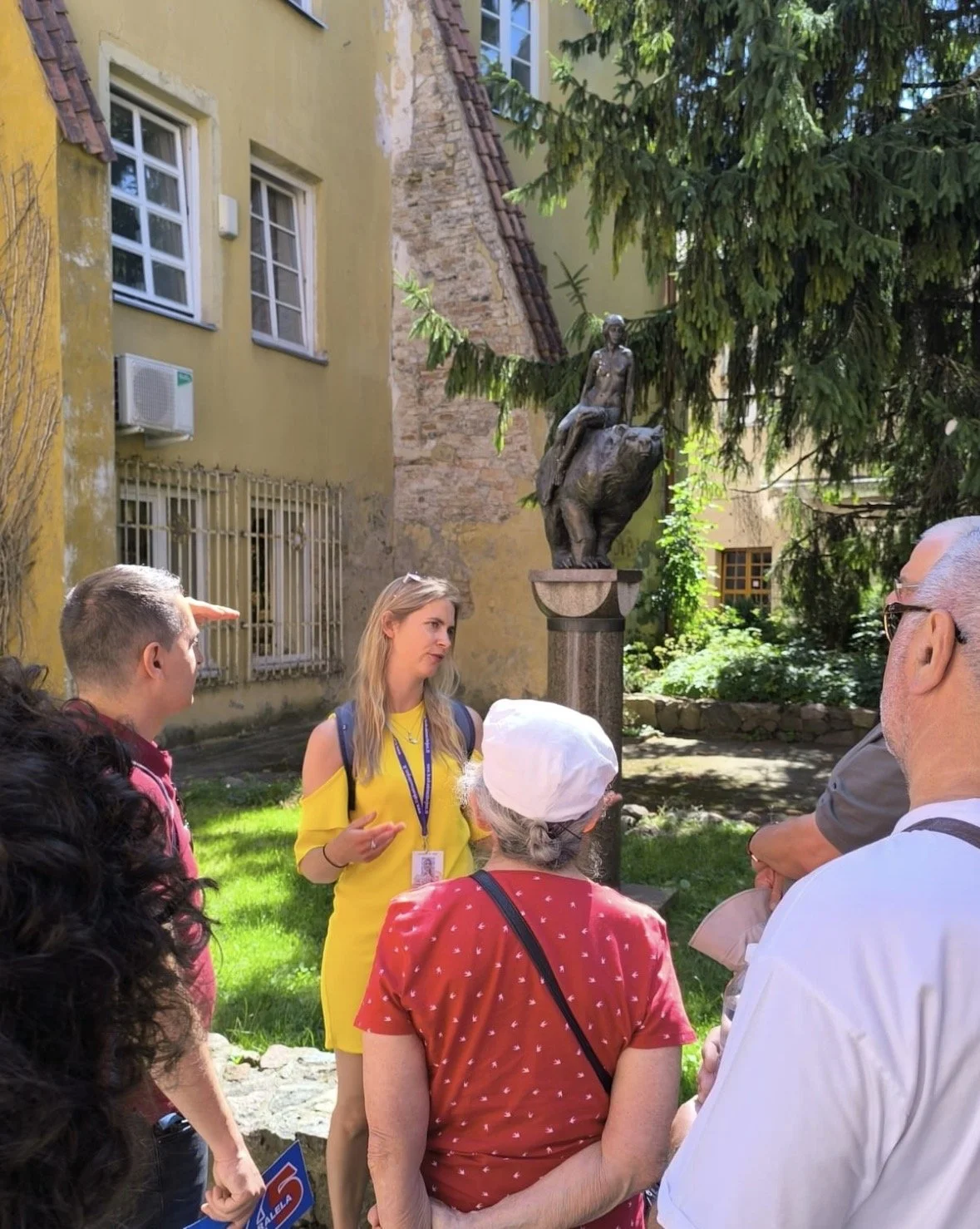 A tour guide explains a sculpture to a group of tourists in an outdoor courtyard surrounded by old buildings and green trees.