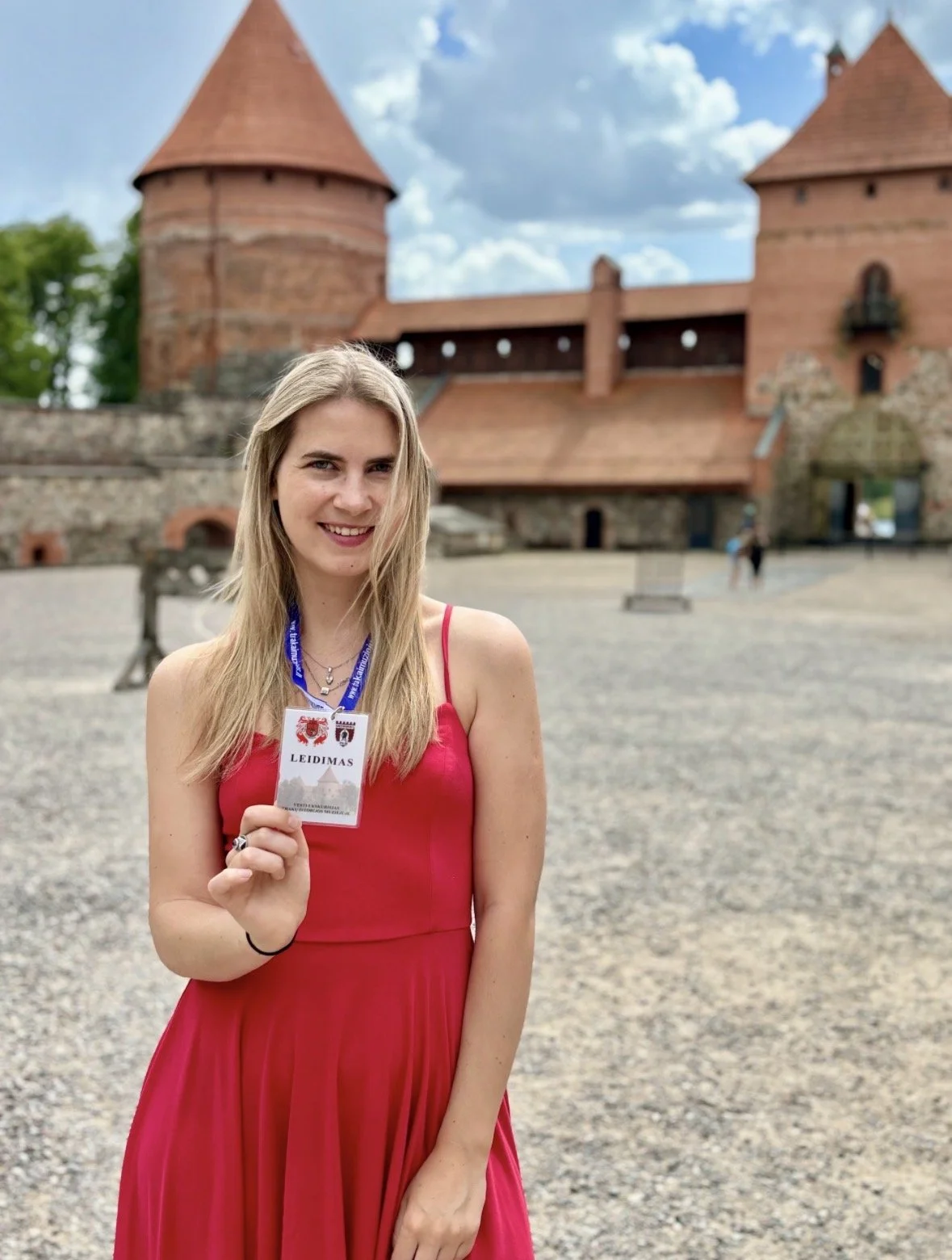 Young woman in a red dress holding an ID badge in front of a historic brick castle with towers.