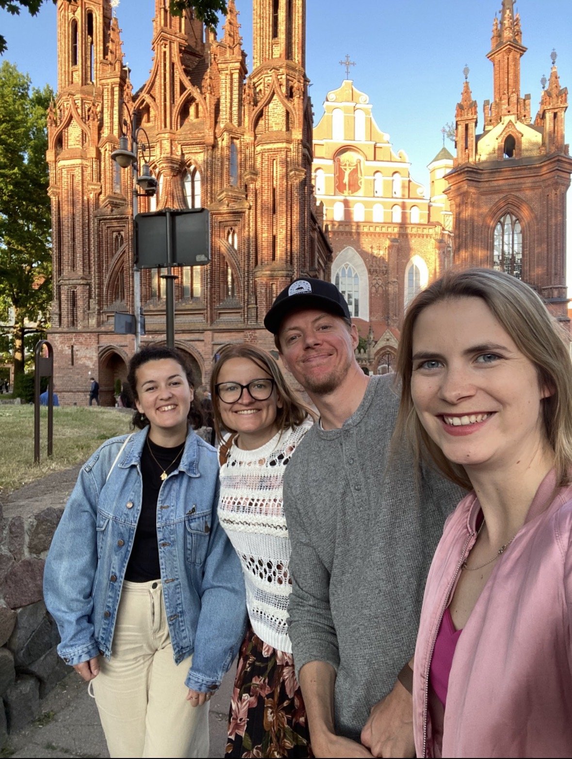 A group of four smiling friends taking a selfie in front of a large, detailed red brick church with tall spires, stained glass windows, and a religious icon on the facade, under a clear blue sky.