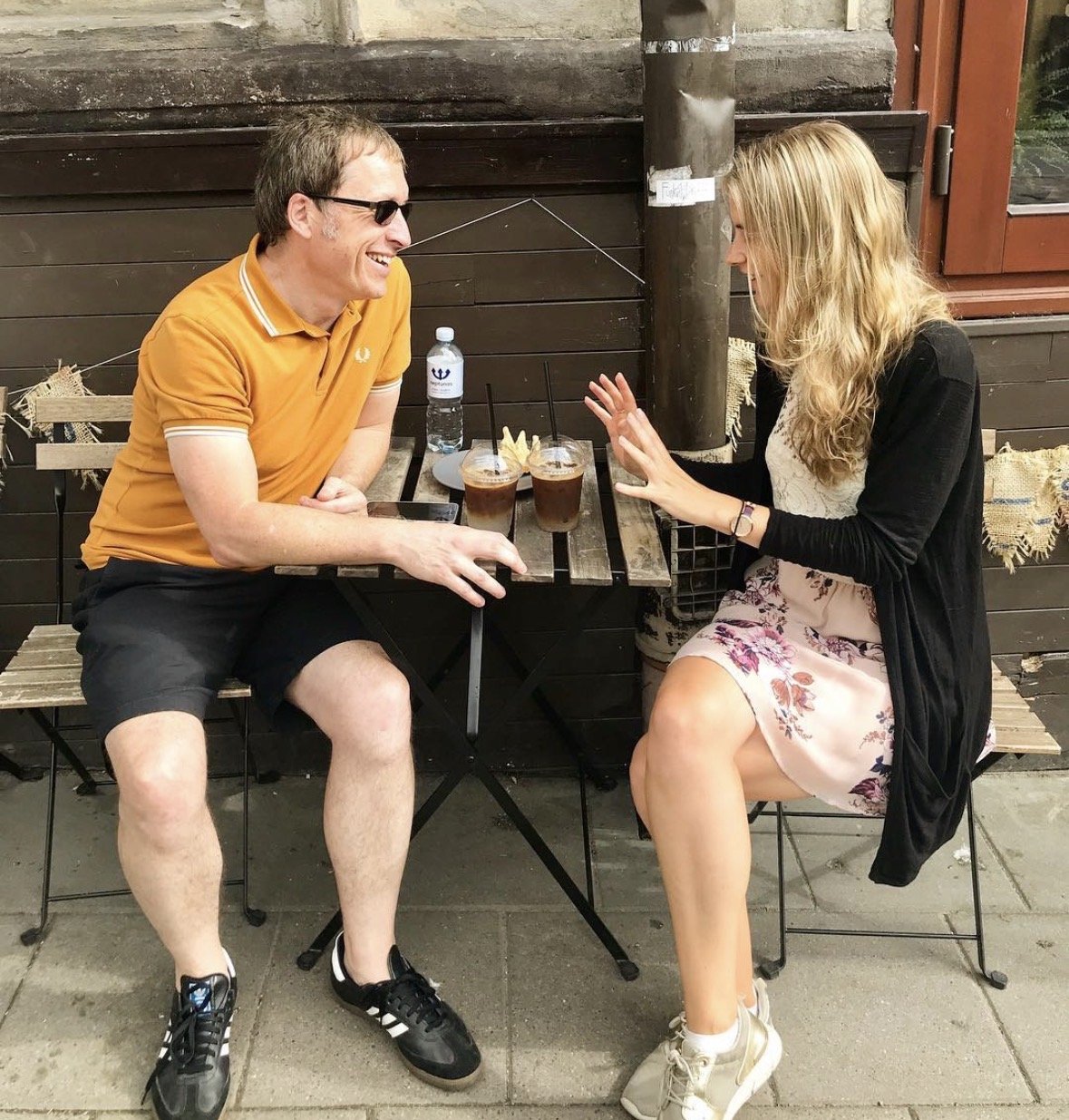 A man and woman sitting at an outdoor table, engaging in conversation, with their drinks on the table. The man is wearing sunglasses and a yellow polo shirt, while the woman has long, wavy blonde hair and is dressed in a floral skirt and black jacket.
