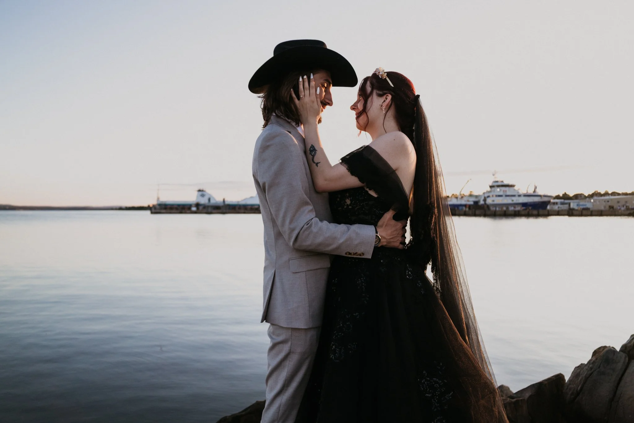 A couple stands close together by a water body at sunset, with ships in the background. The man wears a light suit and wide-brim hat, and the woman wears a black lace dress with an off-the-shoulder design and a headpiece.