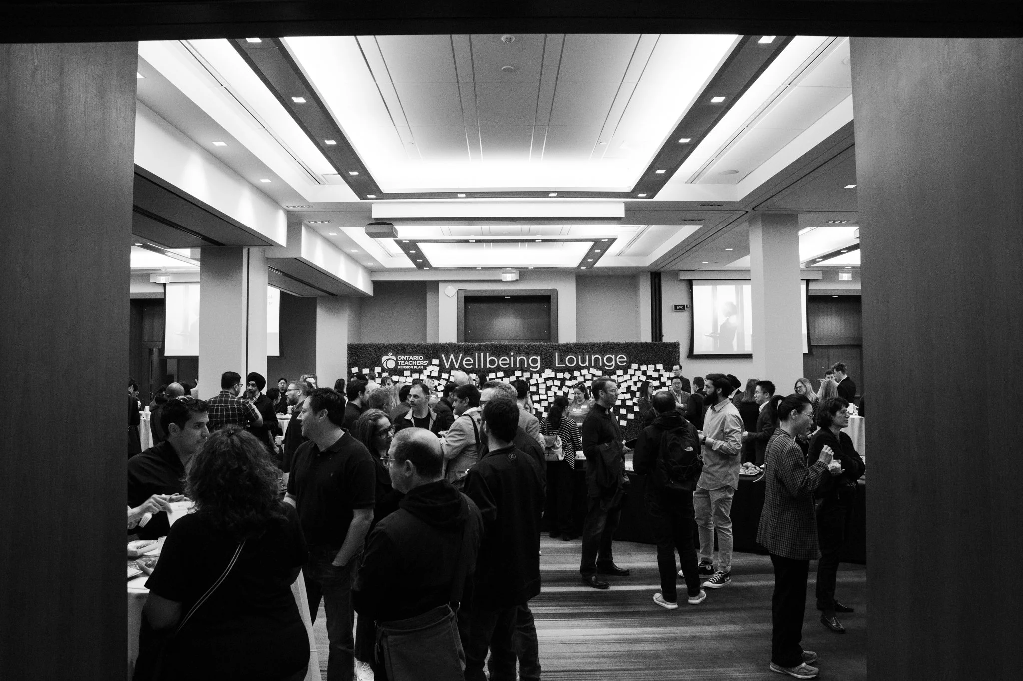 A crowded conference room with people gathered around tables, with a sign that reads 'Wellbeing Lounge' and 'Ontario Teachers' in the background.