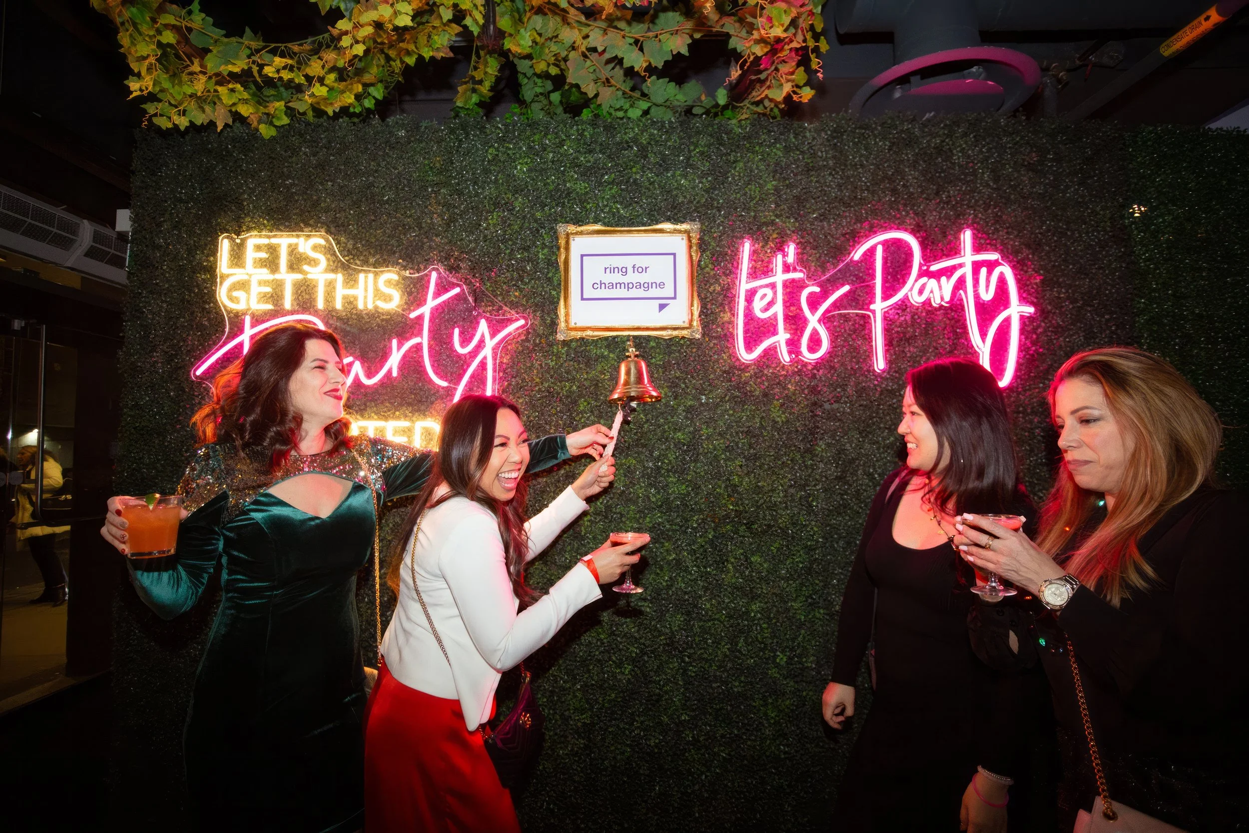 Four women celebrating at a party, holding drinks and ringing a small bell, in front of a neon sign that says 'Let's get this party started' and 'Let's party', with a framed sign that says 'ring for champagne' on a green leafy wall.