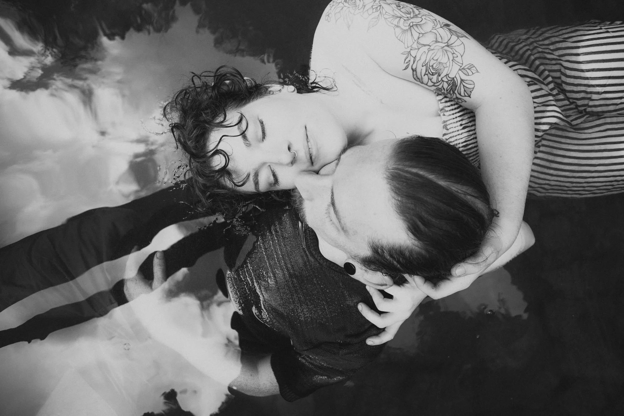 Two women share a kiss and embrace while partially submerged in water, with their faces close together and eyes closed. One woman has curly hair and a septum piercing, and the other has straight hair pulled back. The water creates reflections and a c