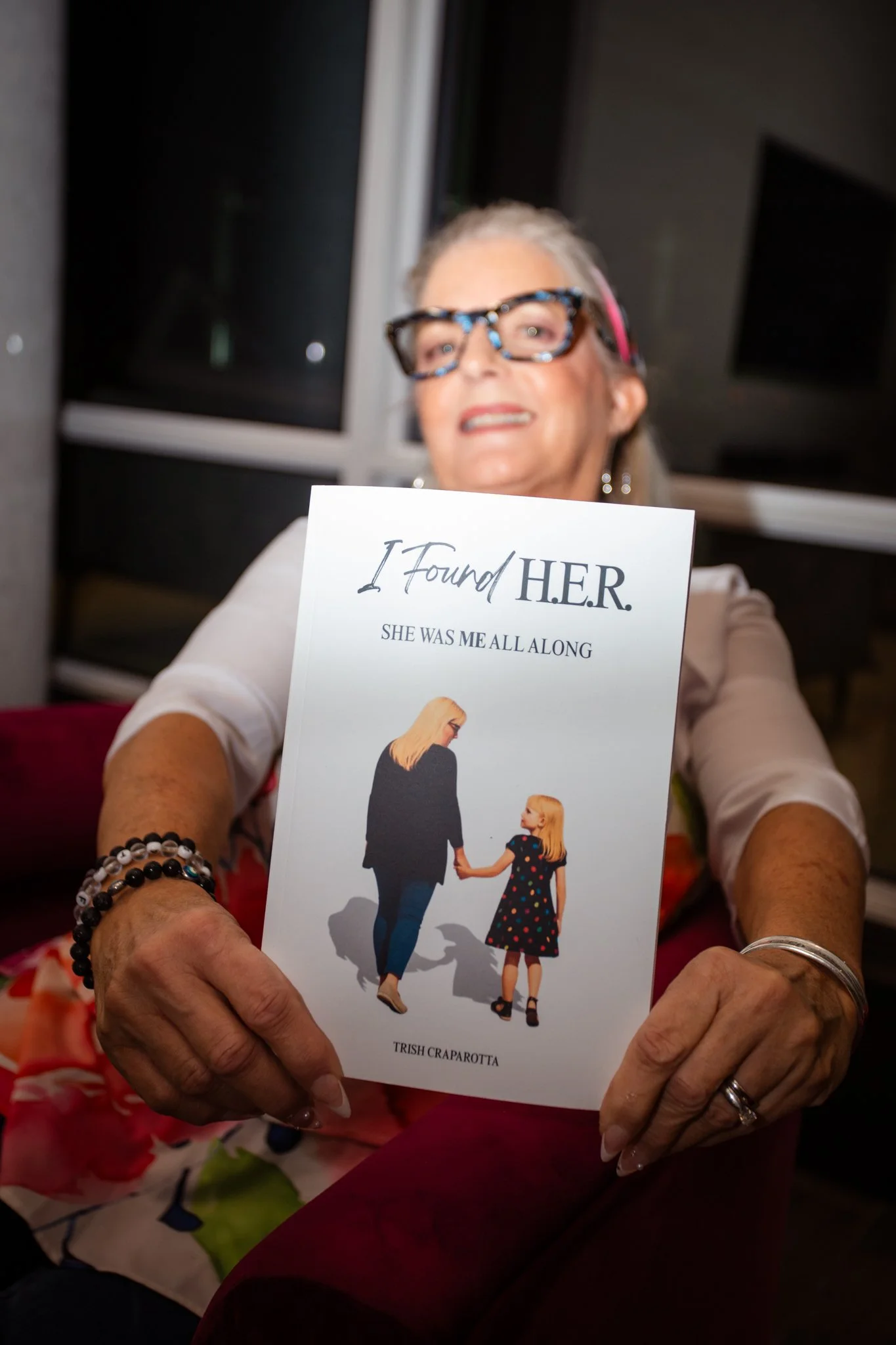 An elderly woman with glasses holding a book titled "I Found HER" featuring a girl and woman holding hands on the cover.