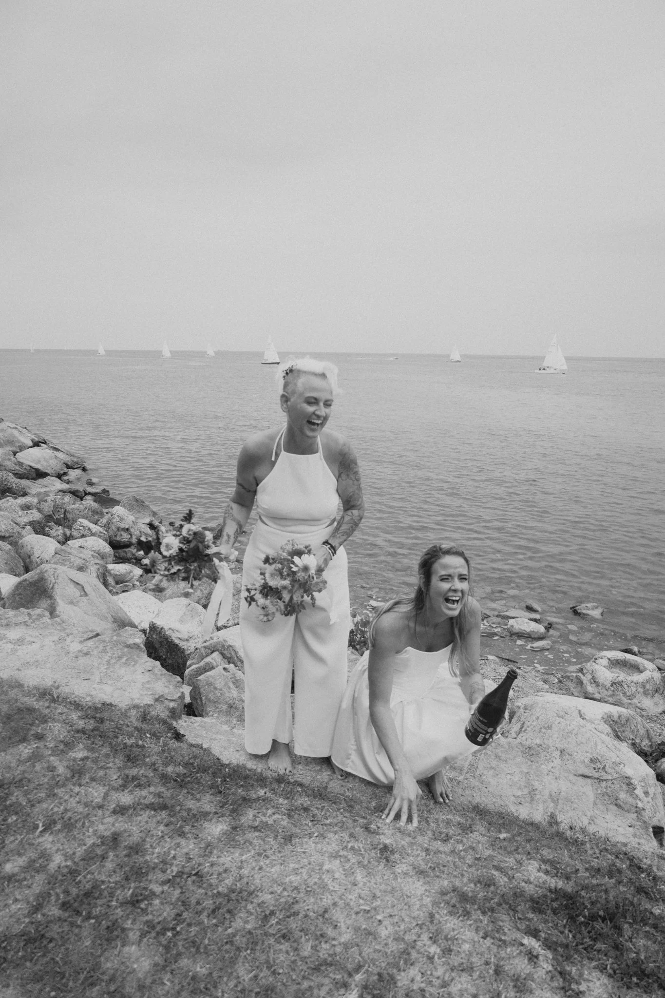 Two women in wedding attire by the water, one standing and holding flowers, the other squatting with a bottle, both laughing and smiling, rocks and sailboats in the background.