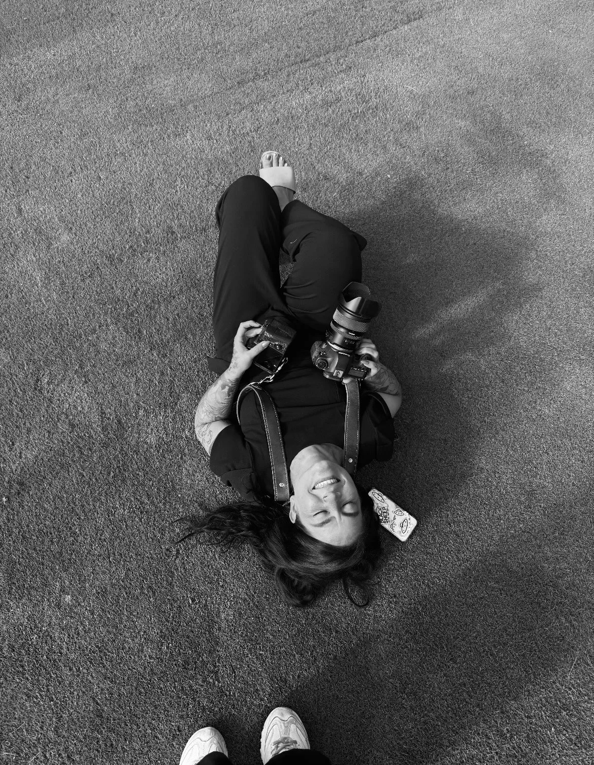 Person lying on the ground with a camera, smiling with eyes closed, and wearing sneakers, seen from above.