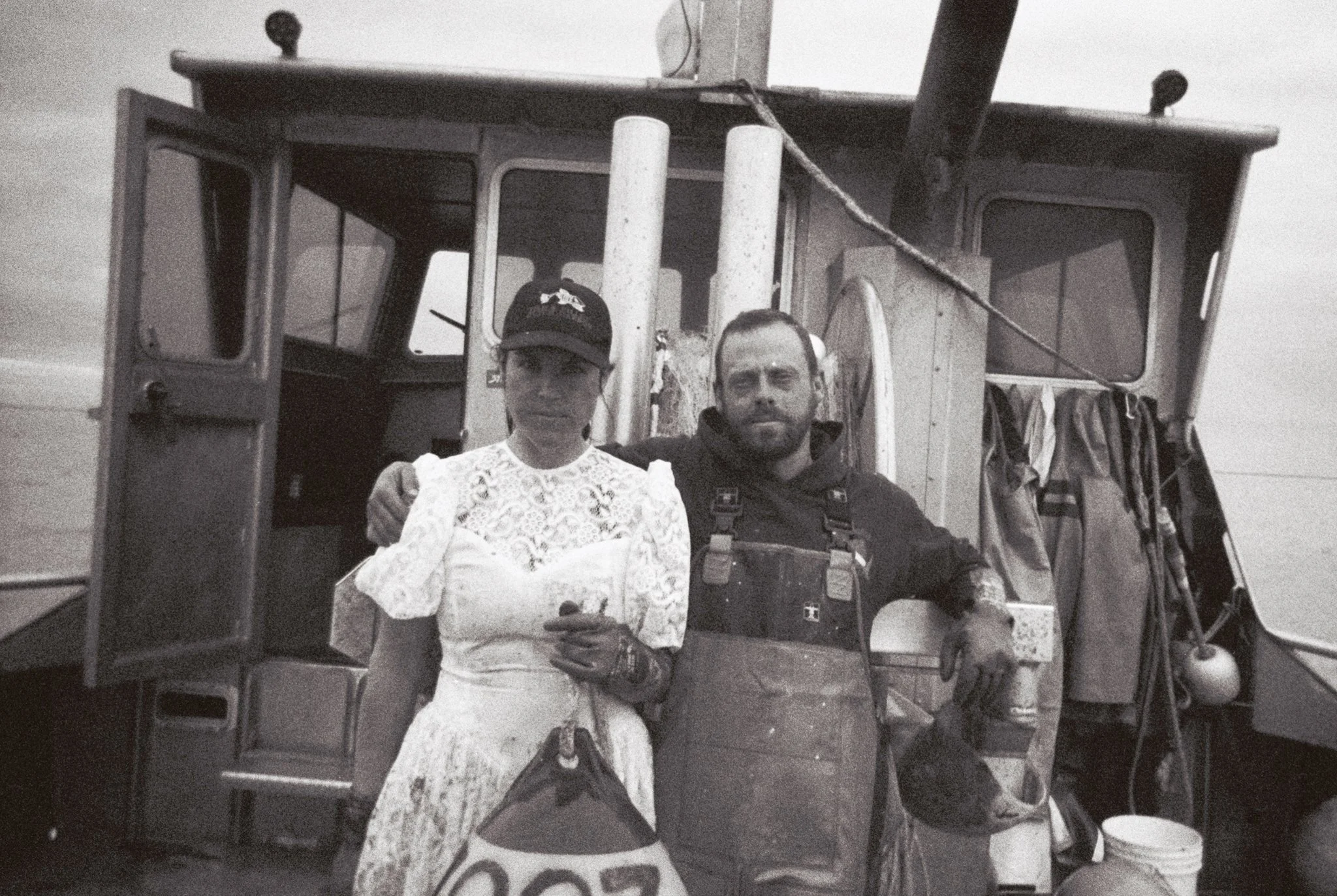 A black-and-white photo of a man and woman standing in front of a vehicle, with the man having his arm around the woman. The woman is wearing a white lace dress and a black cap, while the man is in dark overalls. The vehicle behind them appears to be