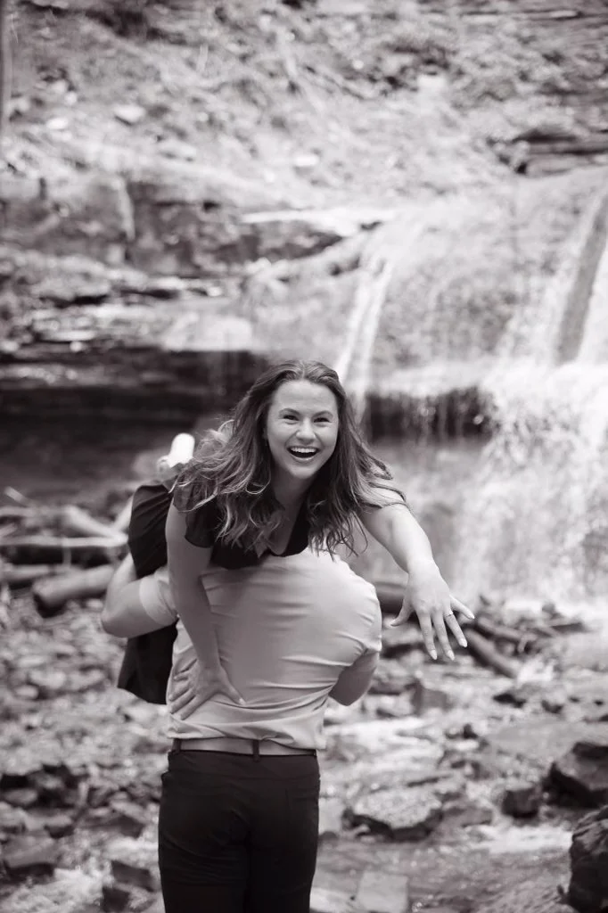 A woman with long hair and a black top has a joyful expression as she is lifted on another person's shoulders in front of a waterfall and rocky landscape.