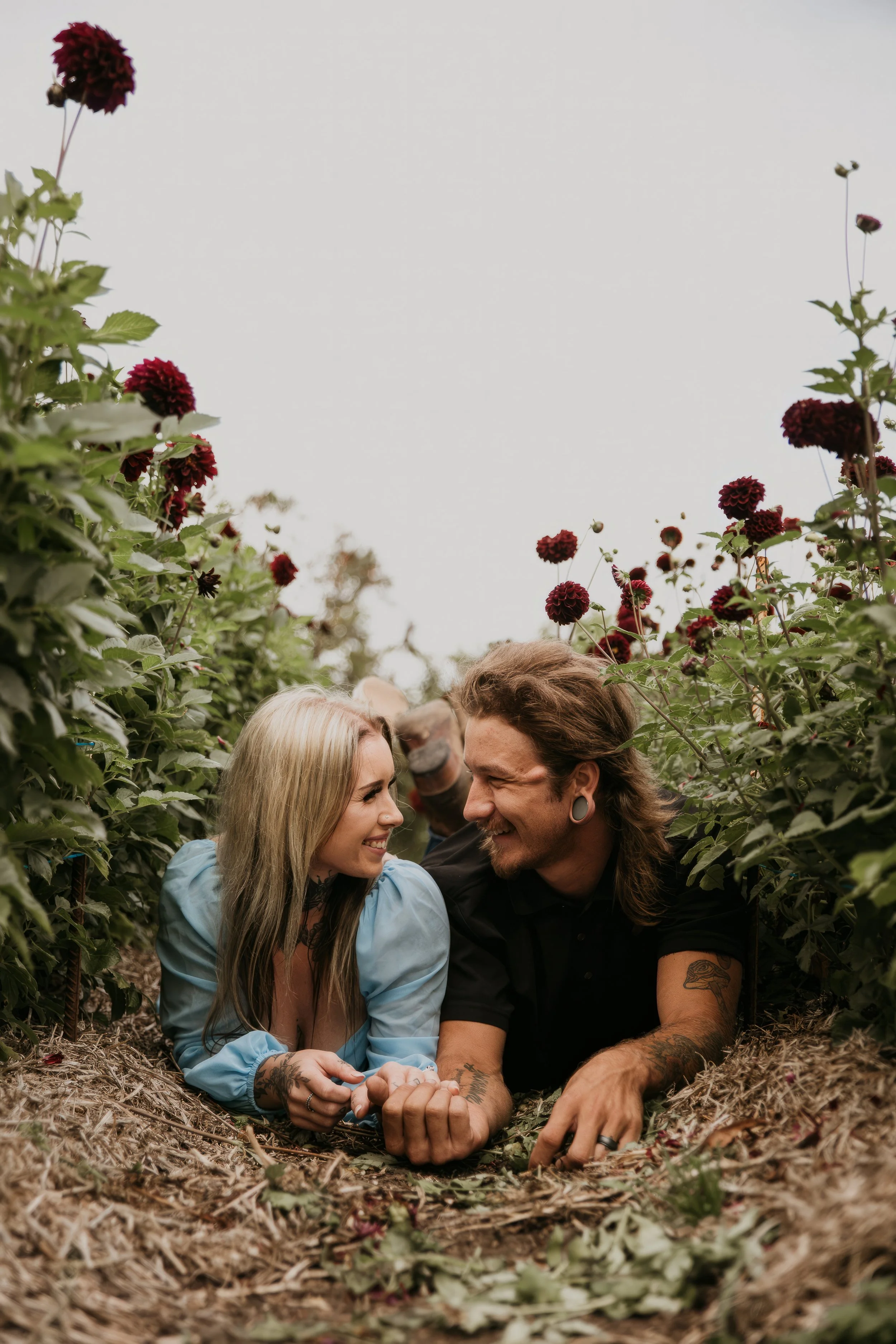 A couple lying on the ground in a flower garden, holding hands and smiling at each other.