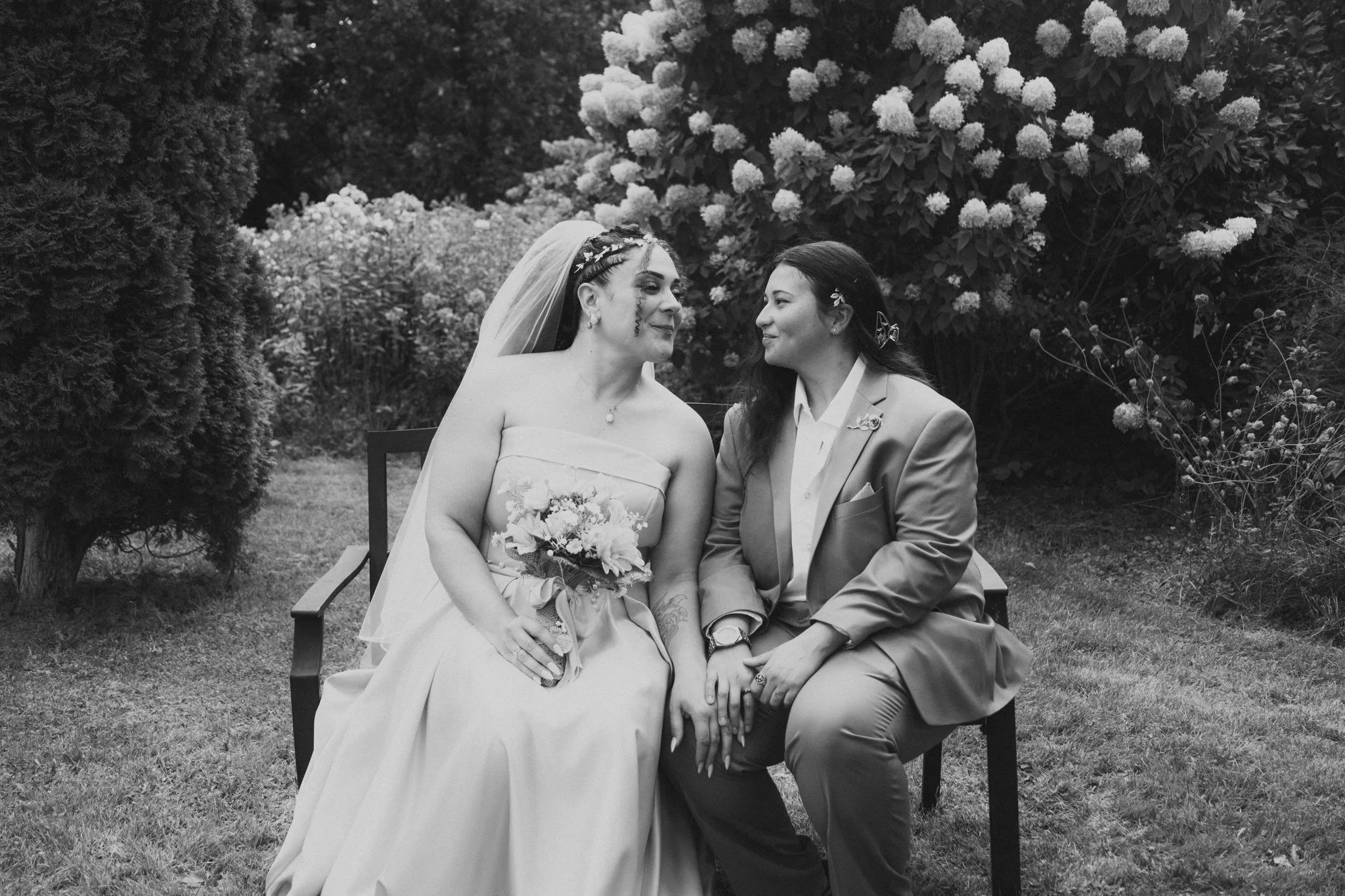 Two women sitting close together on a garden bench, holding hands and looking at each other. The woman on the left is wearing a wedding dress and veil, holding a bouquet, with visible tattoos. The woman on the right is dressed in a suit with a bouton