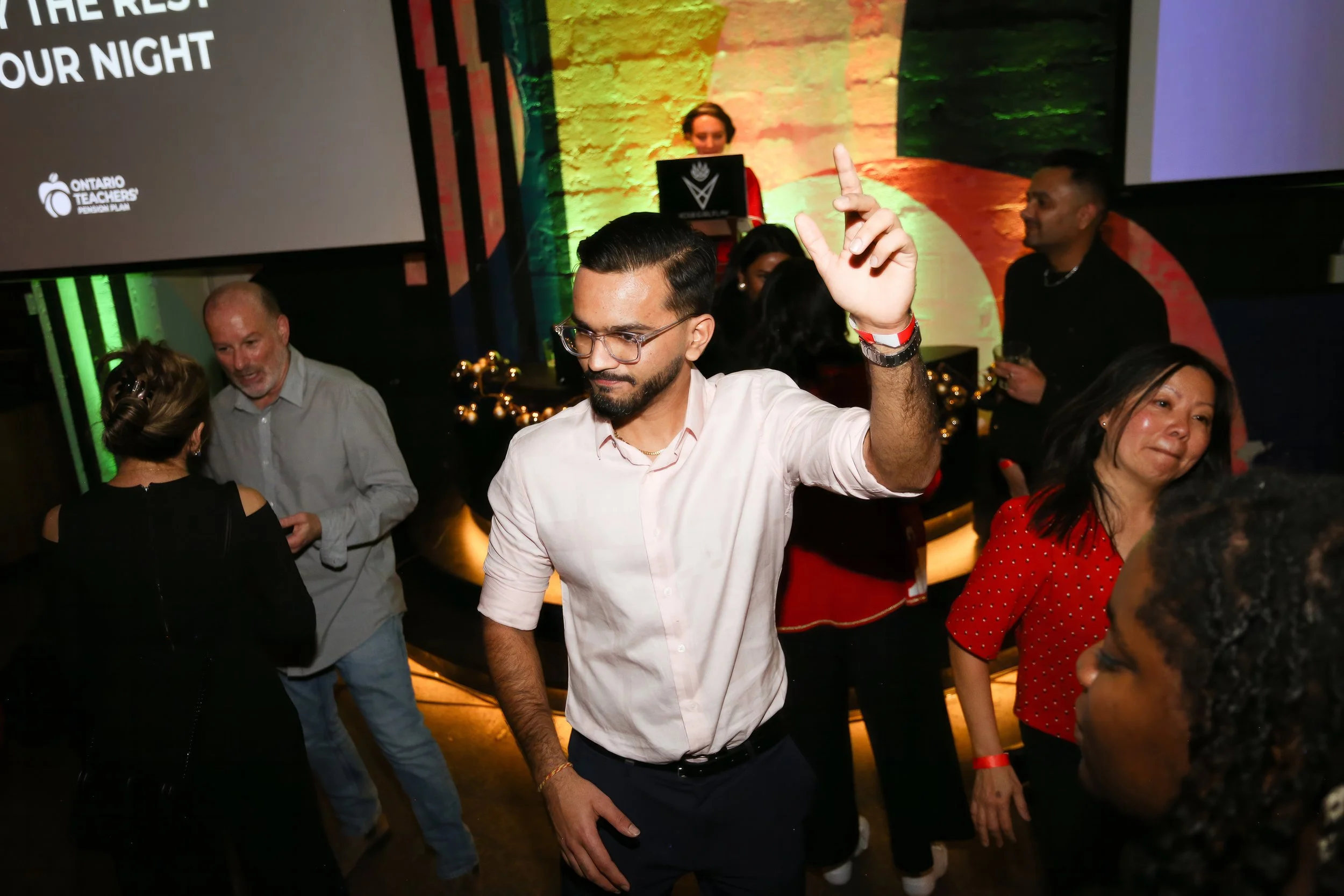 People dancing and socializing at an indoor event with colorful lighting and a stage in the background.