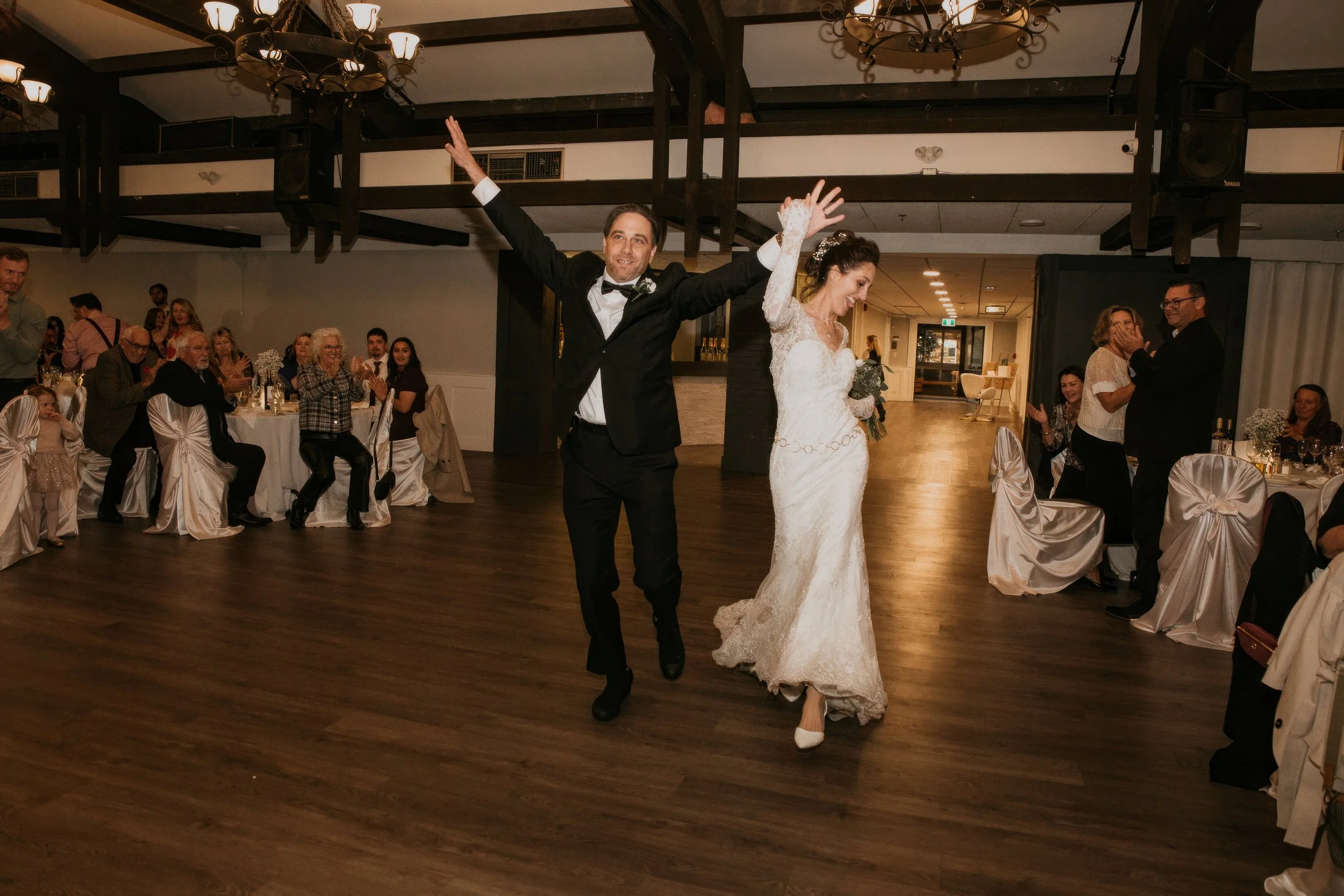 A bride and groom dancing together at their wedding reception, with guests seated at decorated tables clapping and smiling in the background.