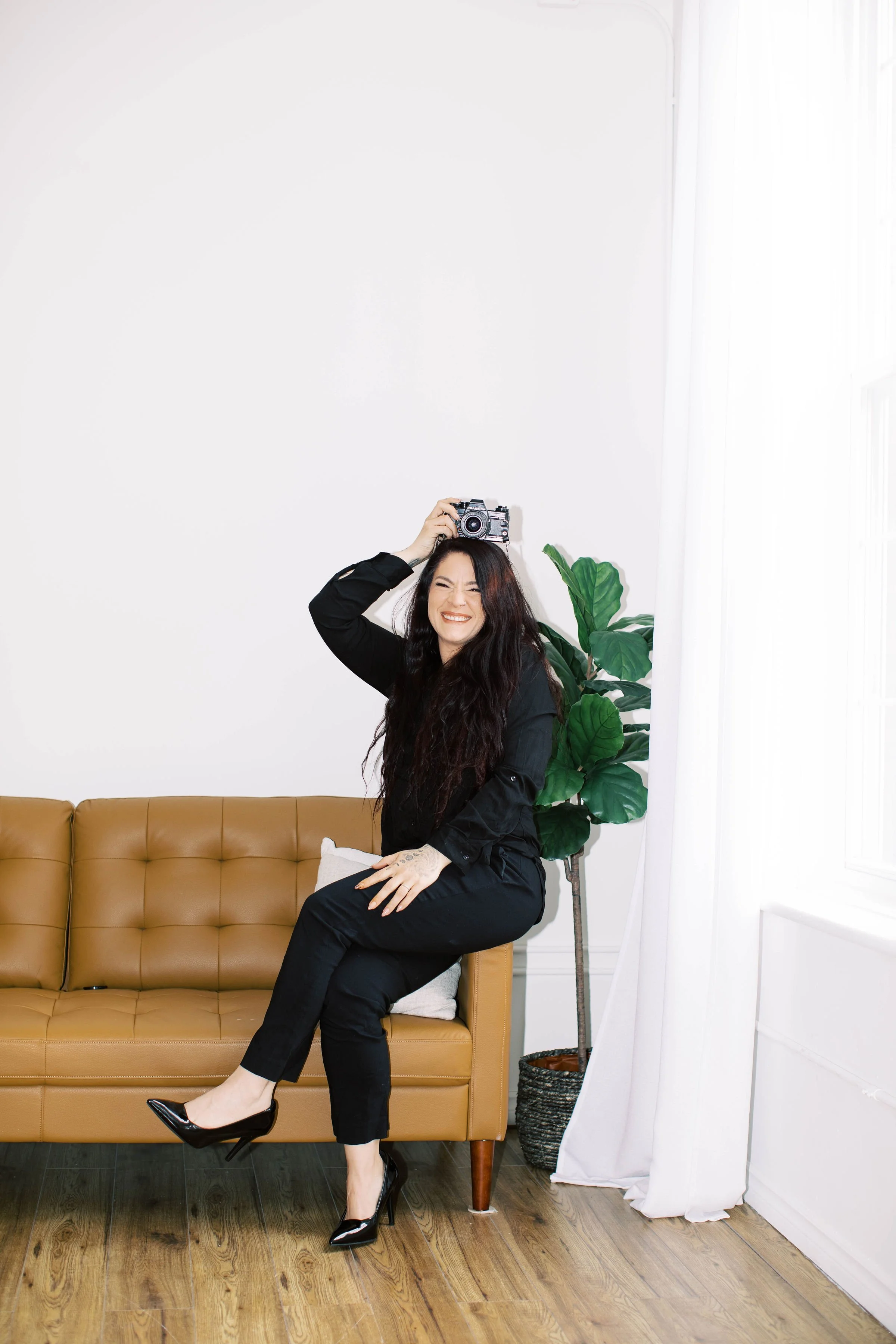 A woman sitting on a tan leather couch, smiling while holding a camera on her head, indoors with a plant and white curtains.
