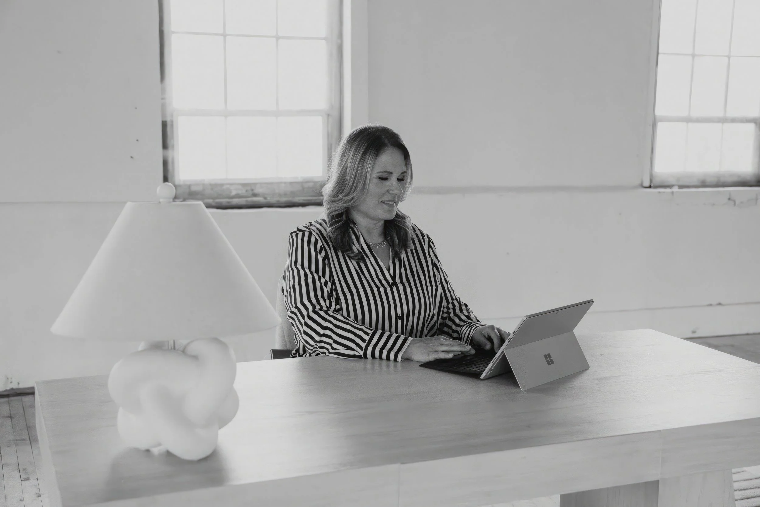 A woman sitting at a wooden table using a tablet device, with a lamp on the table and large windows in the background.