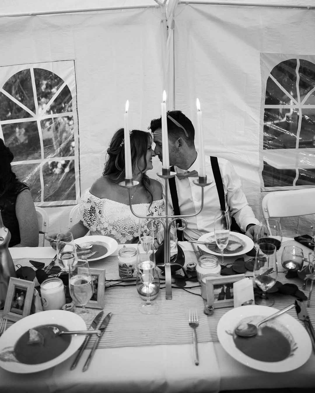 Black and white photo of a bride and groom sitting at a decorated dining table inside a tent, leaning their foreheads together near a candelabra with lit candles, surrounded by dinnerware and decorative elements.