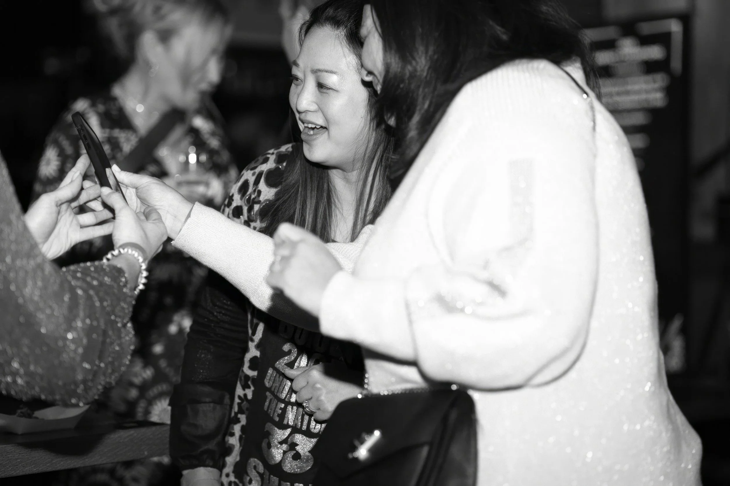 Several women are gathered together, looking at a phone held by one of them, smiling and engaging in conversation.