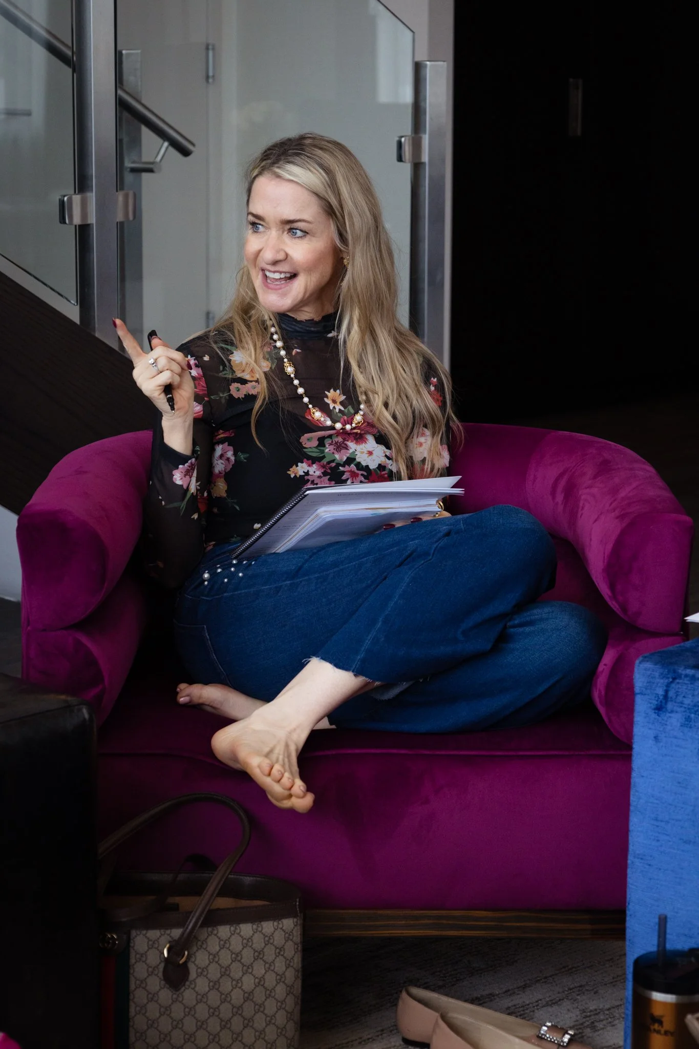 A woman with long blonde hair, wearing a floral patterned black top, blue jeans, sits cross-legged on a magenta couch. She is smiling and gesturing with her right hand, holding a pen, with papers on her lap. The background shows a glass door and a da