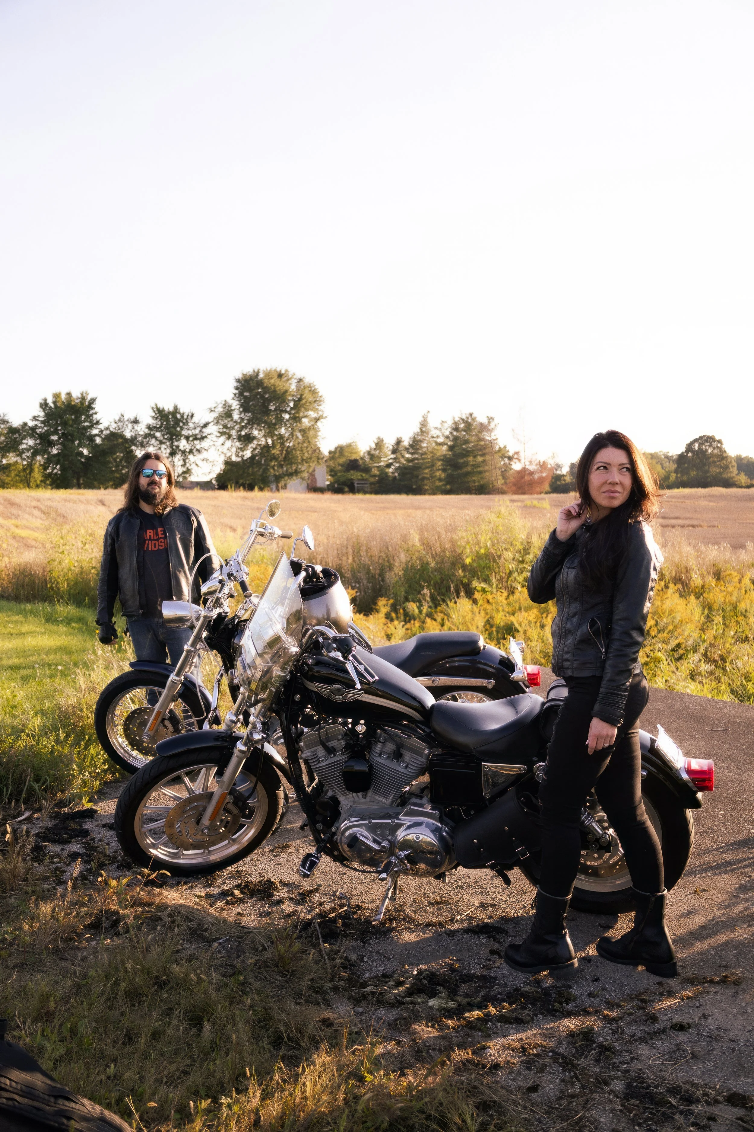 Two motorcyclists, a man with long hair and a woman with dark hair, standing on a rural road with motorcycles during sunset. The man is wearing a black leather jacket, sunglasses, and denim jeans, while the woman is dressed in a black leather jacket,