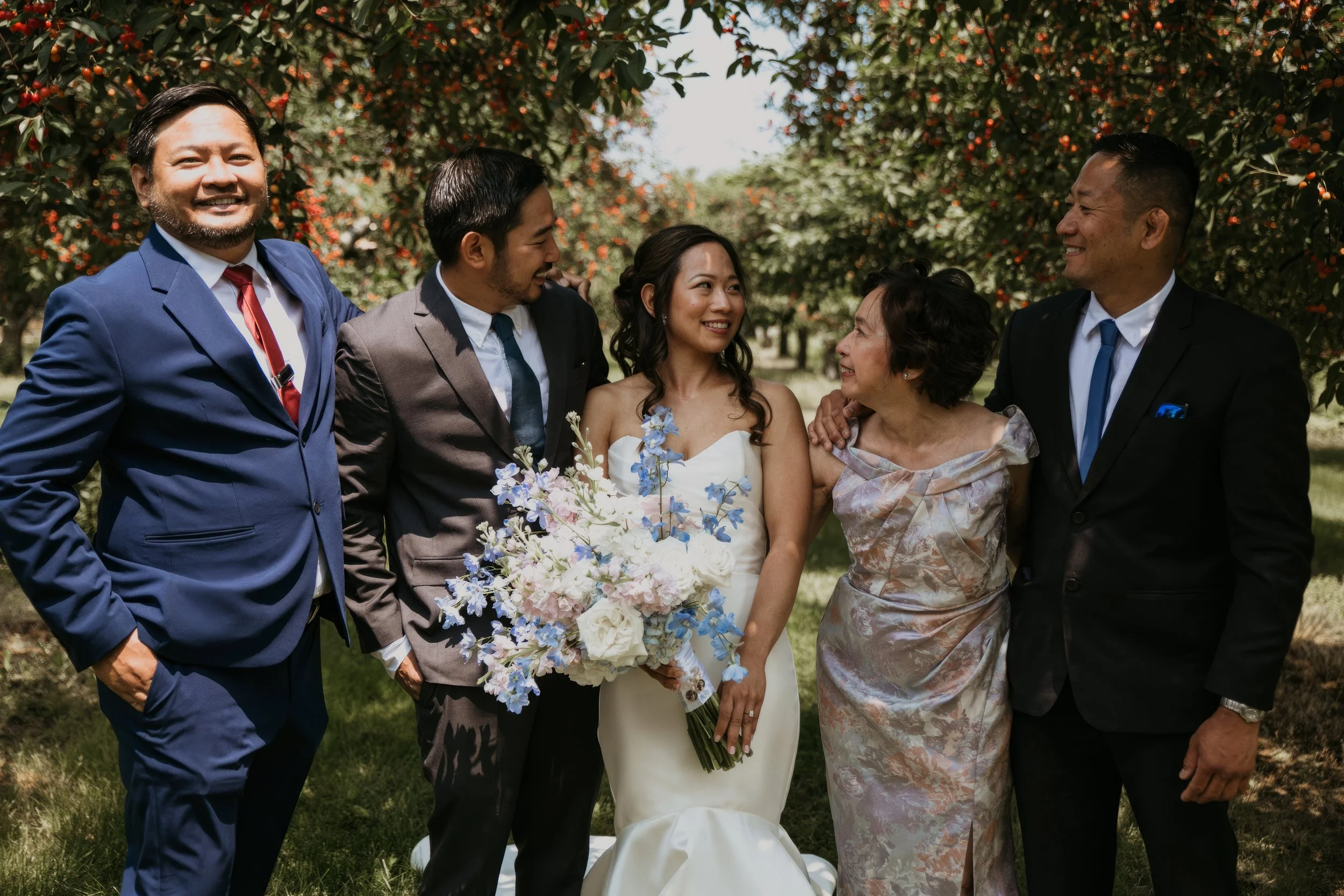 Group of six people at a wedding outdoors, including bride in a white wedding dress holding a bouquet of flowers, surrounded by family and friends, smiling and enjoying the moment under trees with orange fruit.