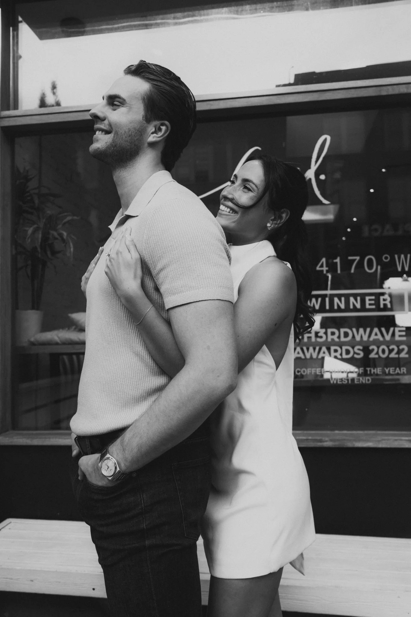 A smiling woman hugging a man from behind outside a building with a sign that reads 'Winner' and '2022 Hardwave Awards'.