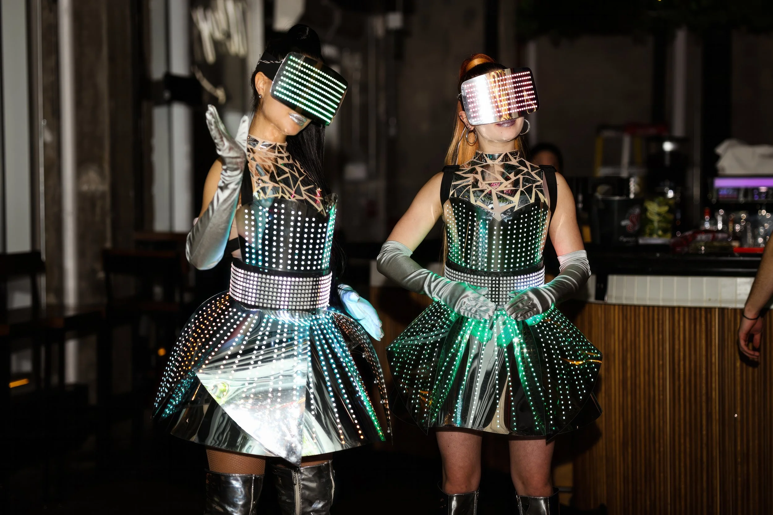 Two women wearing futuristic, reflective LED-lit outfits and LED masks, standing indoors at a party or event.